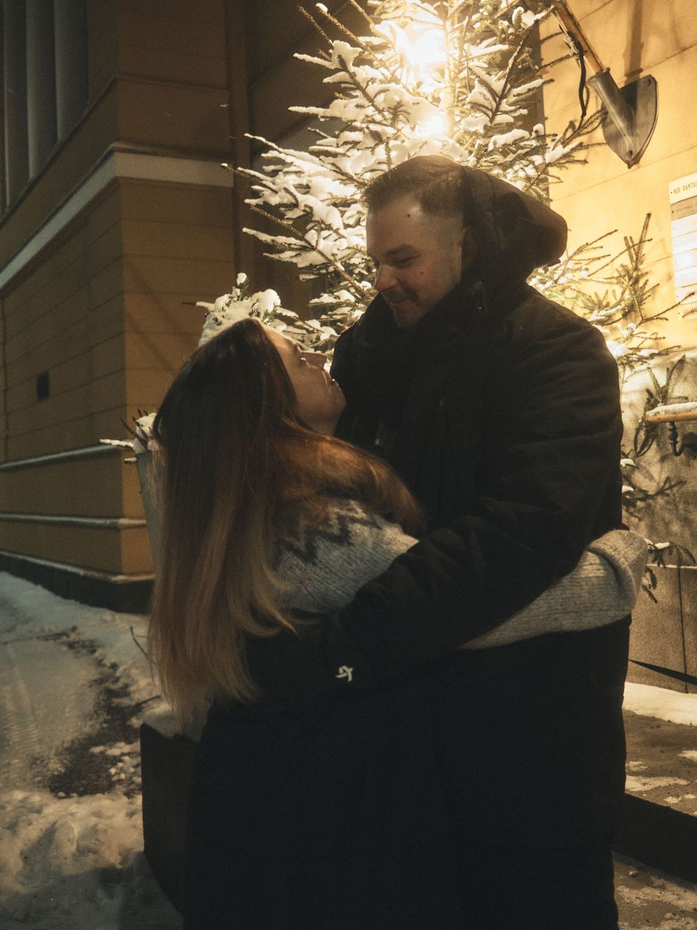 A couple embraces and kisses outside in the snow at night with warm lighting creating a romantic winter atmosphere.