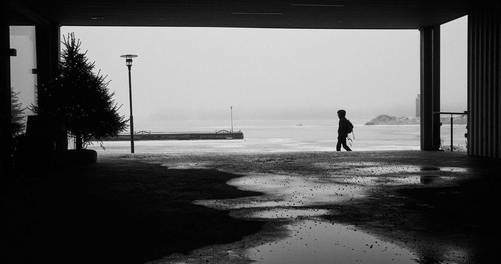 A silhouetted figure walks through a covered walkway on a misty day, creating a dramatic black and white composition.