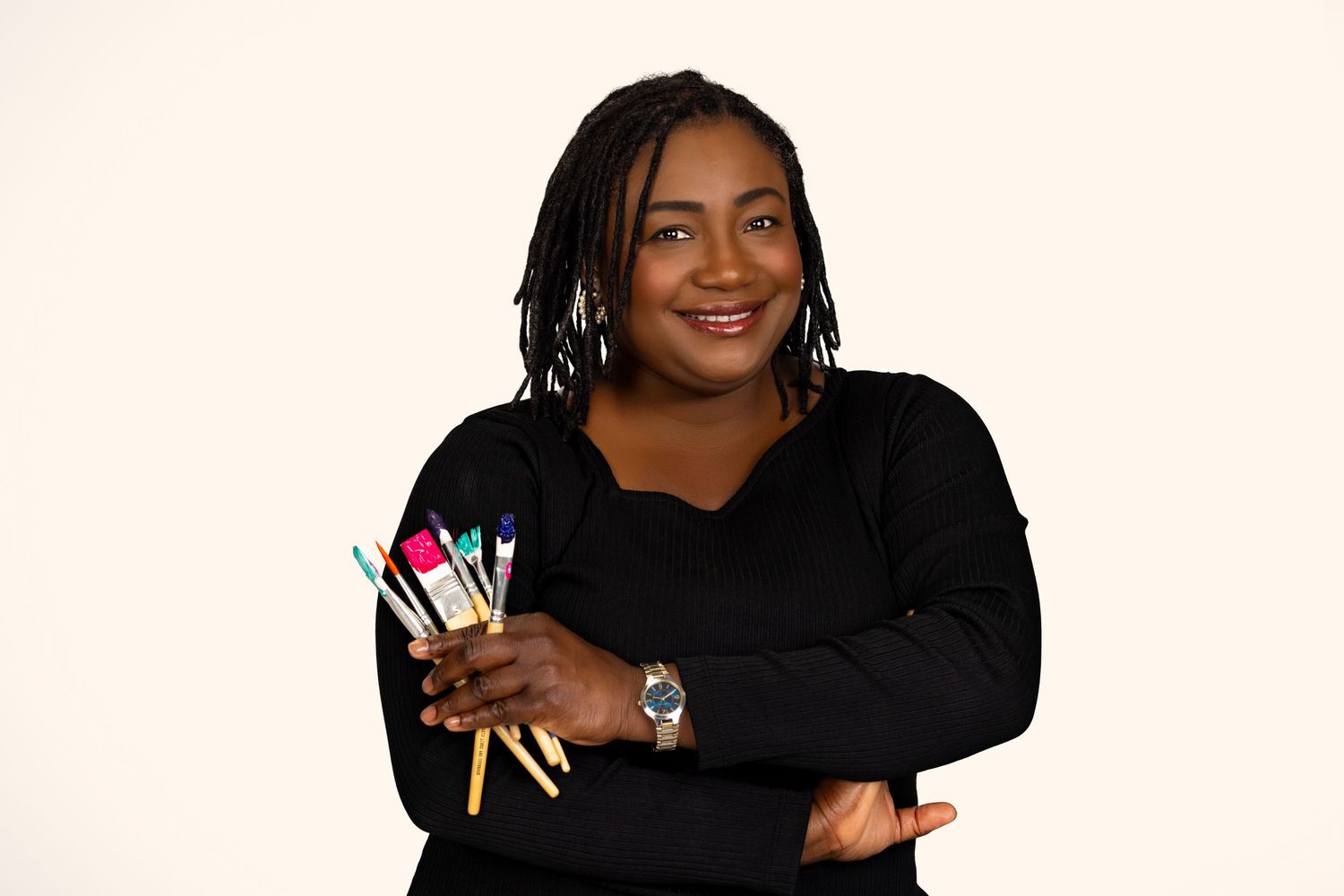 A smiling woman holds colorful paintbrushes while posing against a white background.