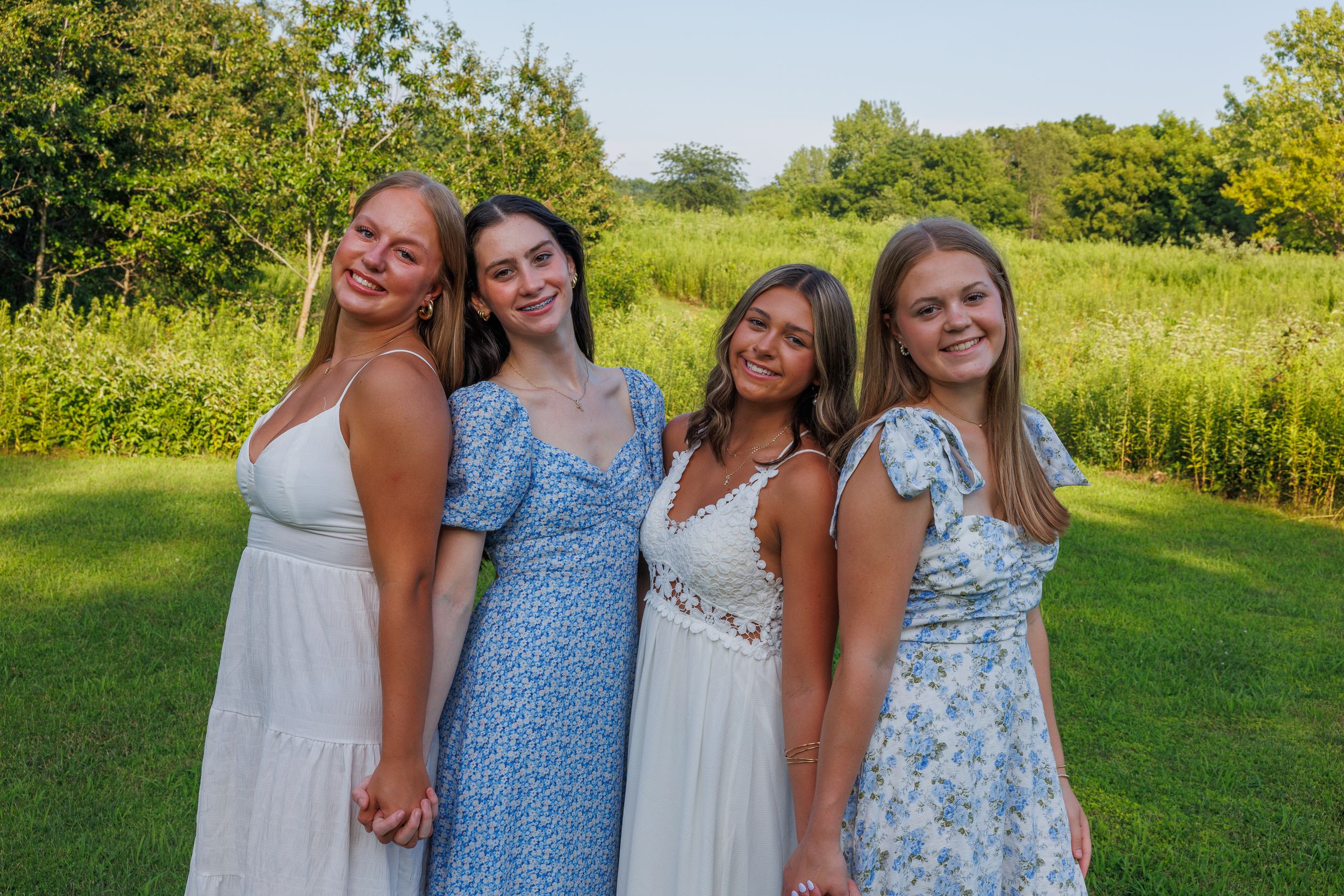 A group in blue and white summer dresses poses together in a sunny green field with wildflowers.