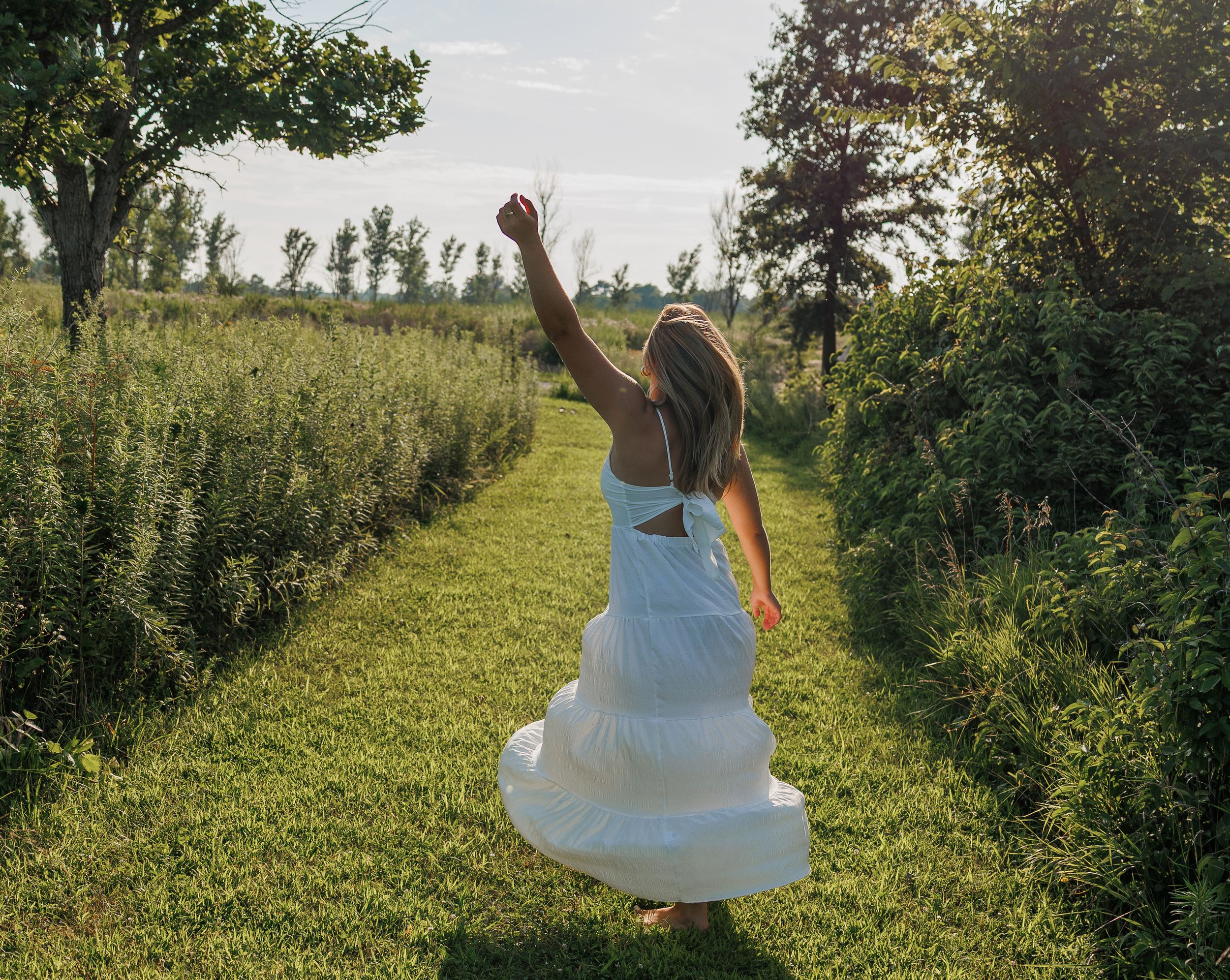 Someone in a flowing white dress twirls joyfully in a sunlit meadow surrounded by green trees and grass.