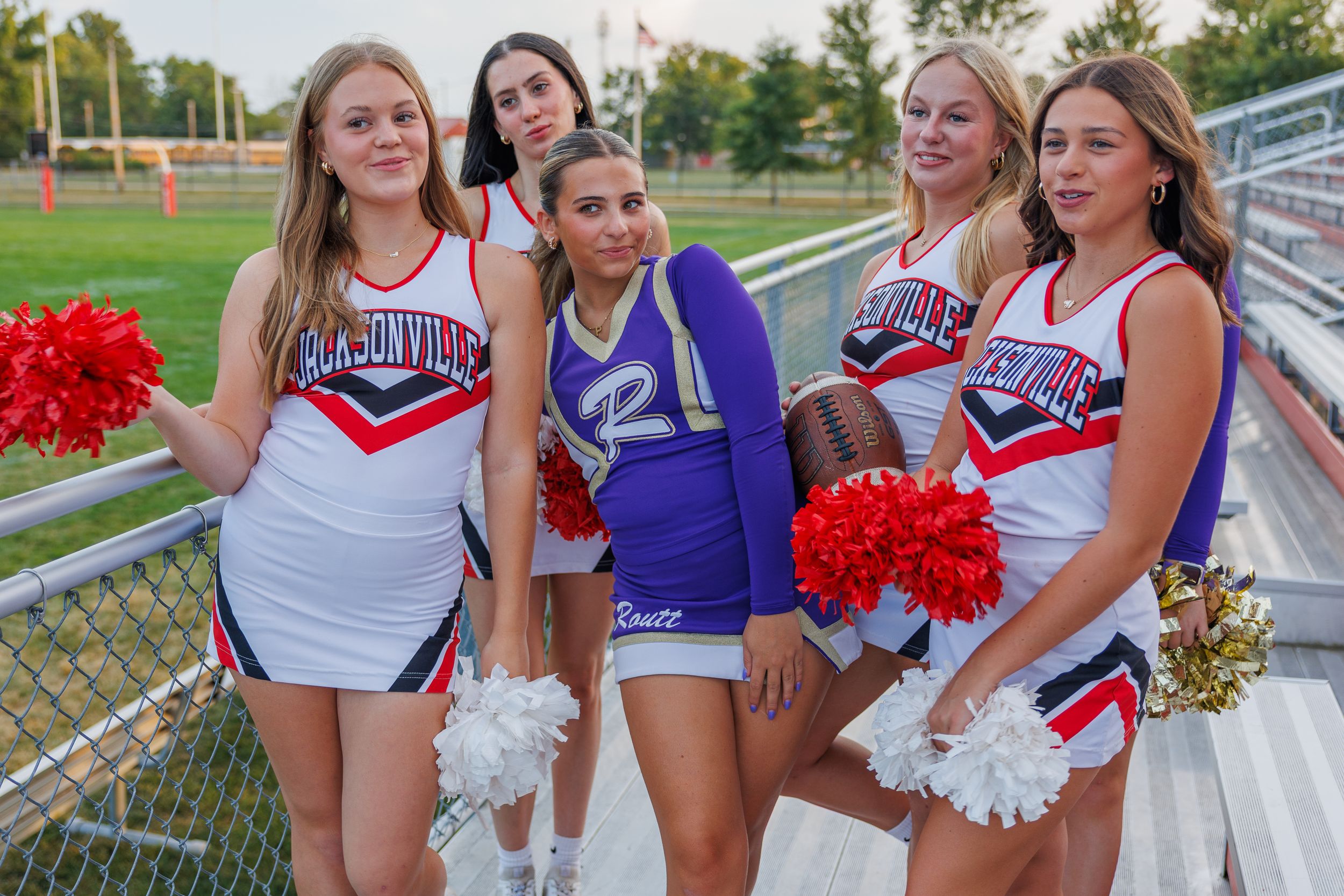 High school cheerleaders in red white and blue uniforms pose with pom poms on bleachers at an outdoor sports field.