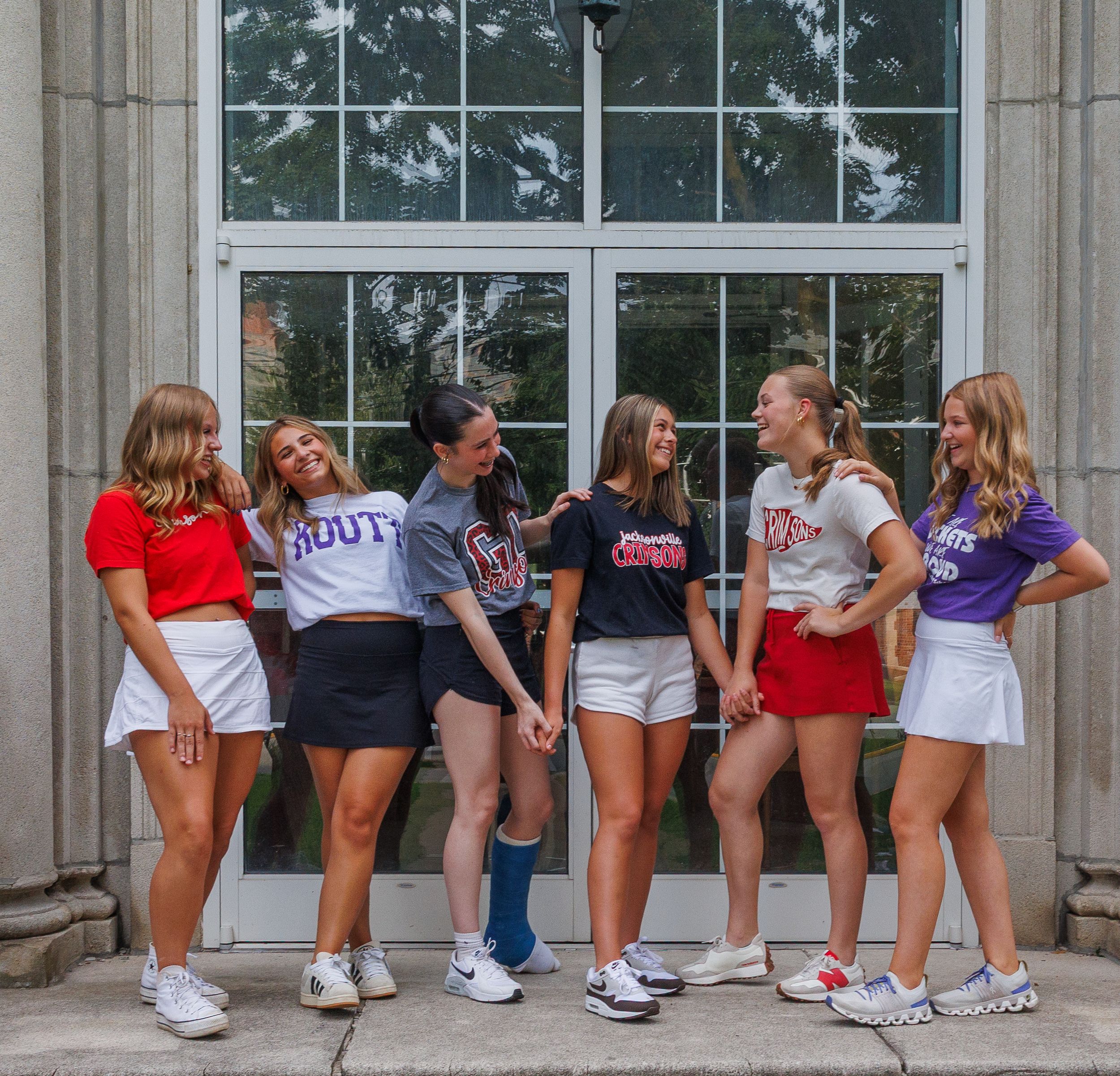 Group of students in collegiate apparel and athletic shoes pose together on stone steps outside a building window.