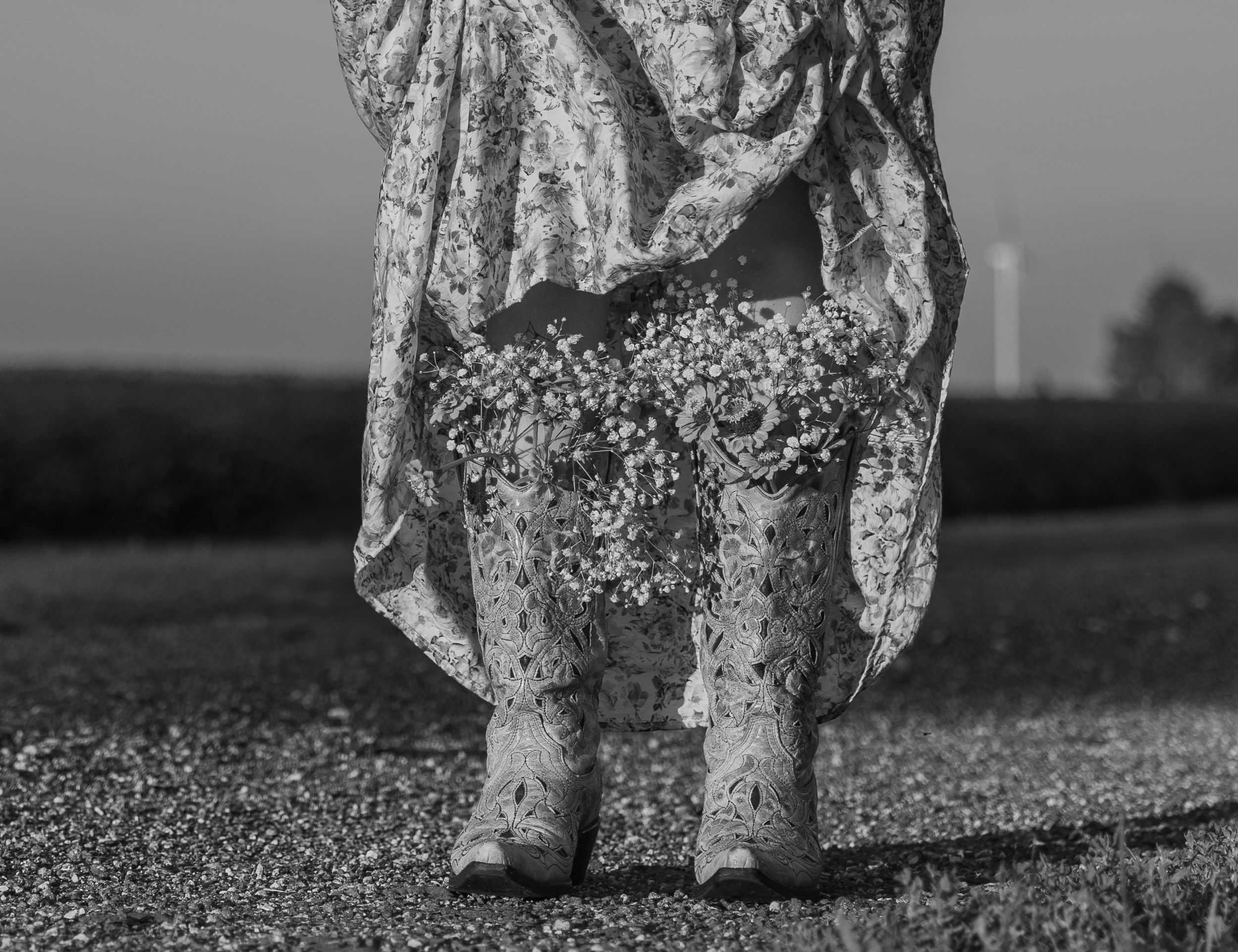 Black and white close-up of feet walking through grass in vintage floral dress and boots.