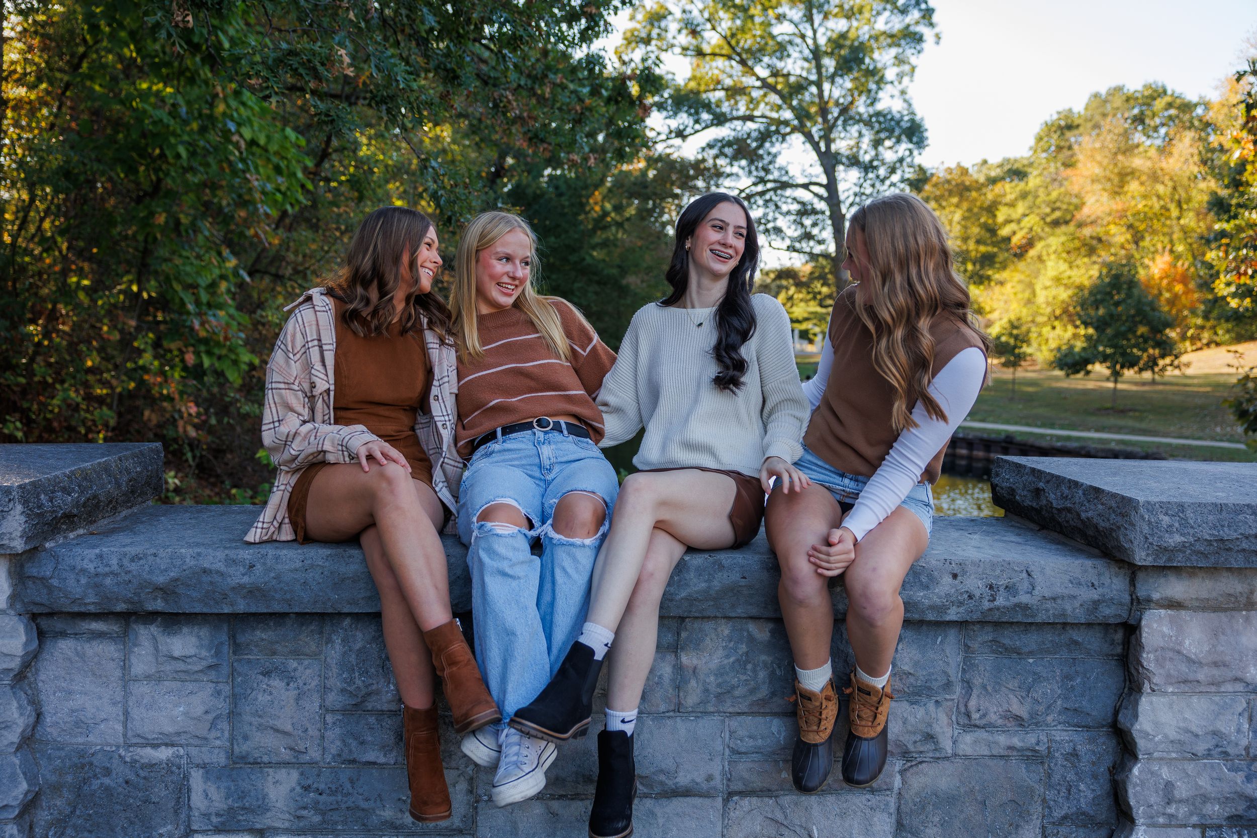 Four friends in casual fall outfits sitting together on a stone wall on a sunny autumn afternoon.