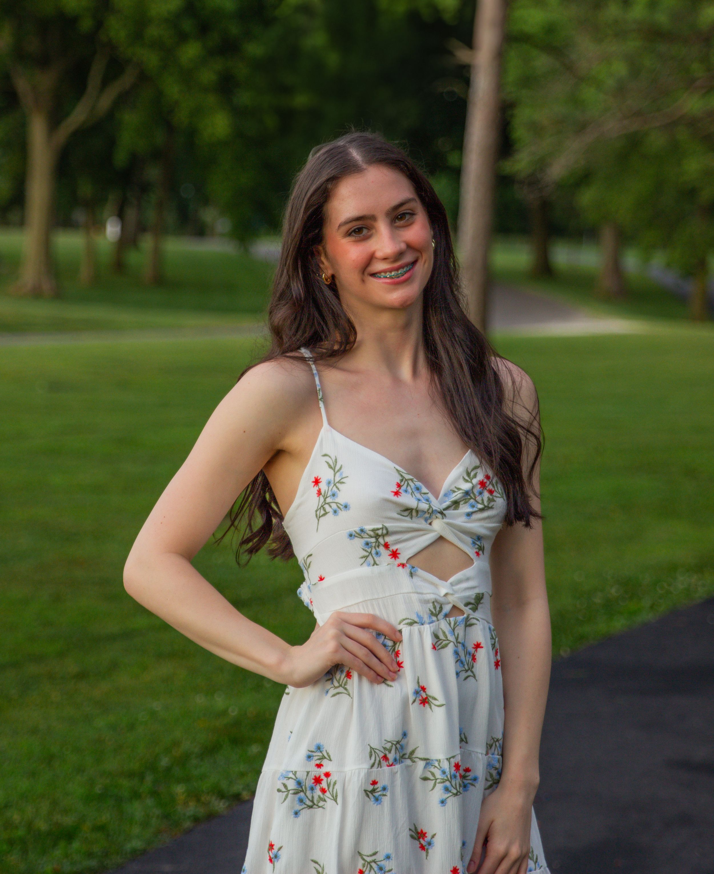 Woman in white floral sundress smiling outdoors in park setting with green trees in background.