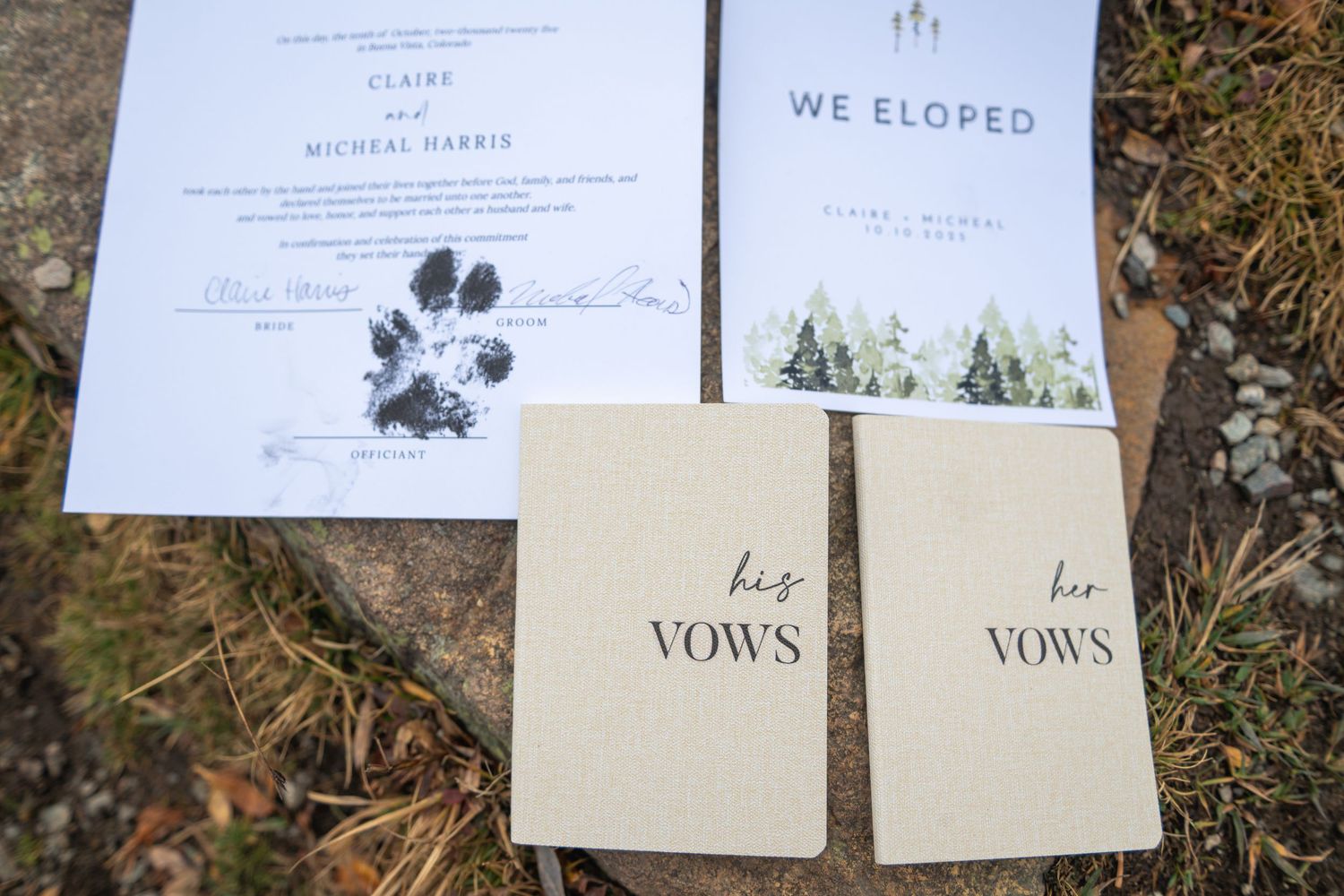 Wedding vow books and a marriage license with paw print detail lay on natural outdoor ground.