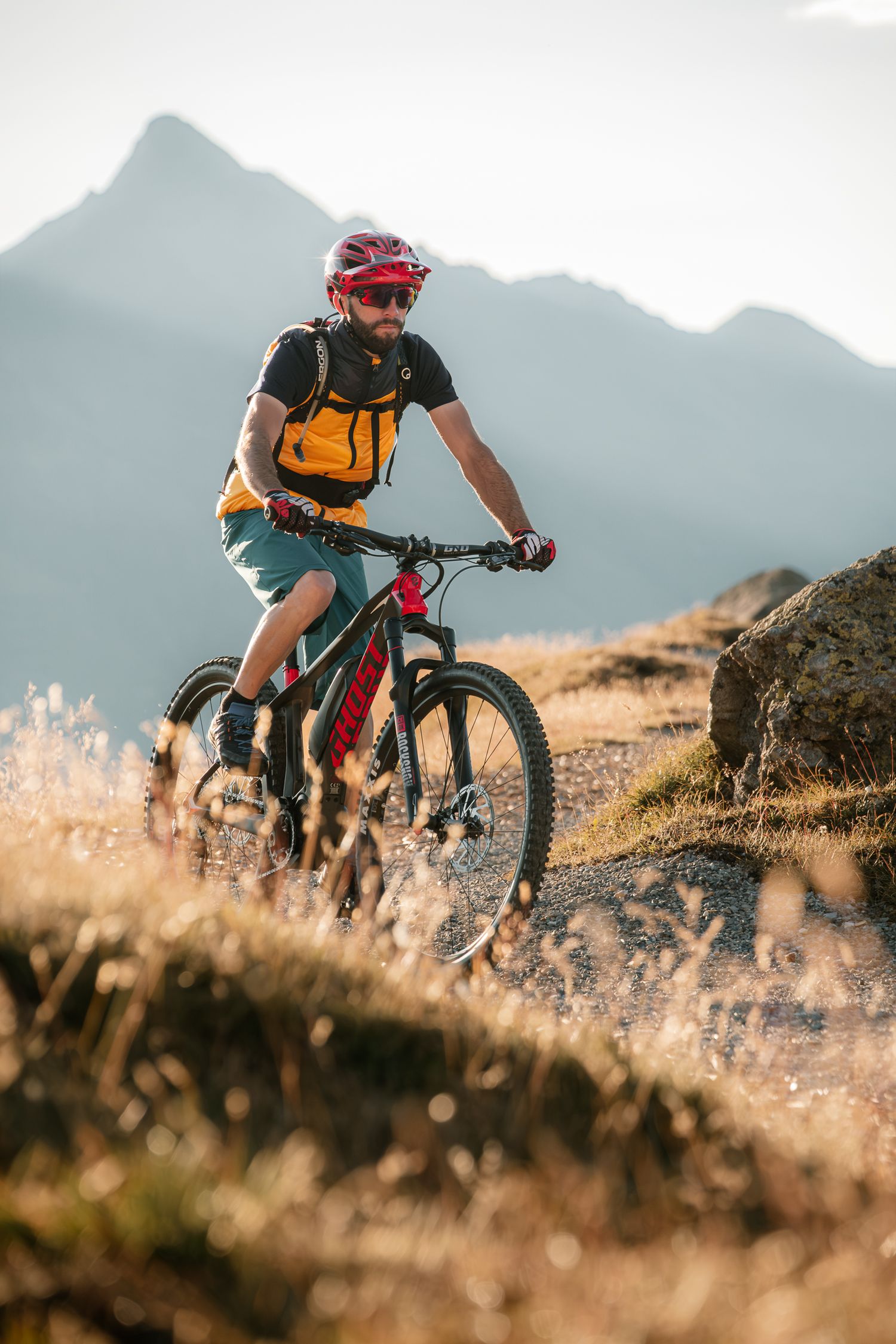 Mountain biker rides trail at sunset with orange backpack and red helmet against misty mountain backdrop.
