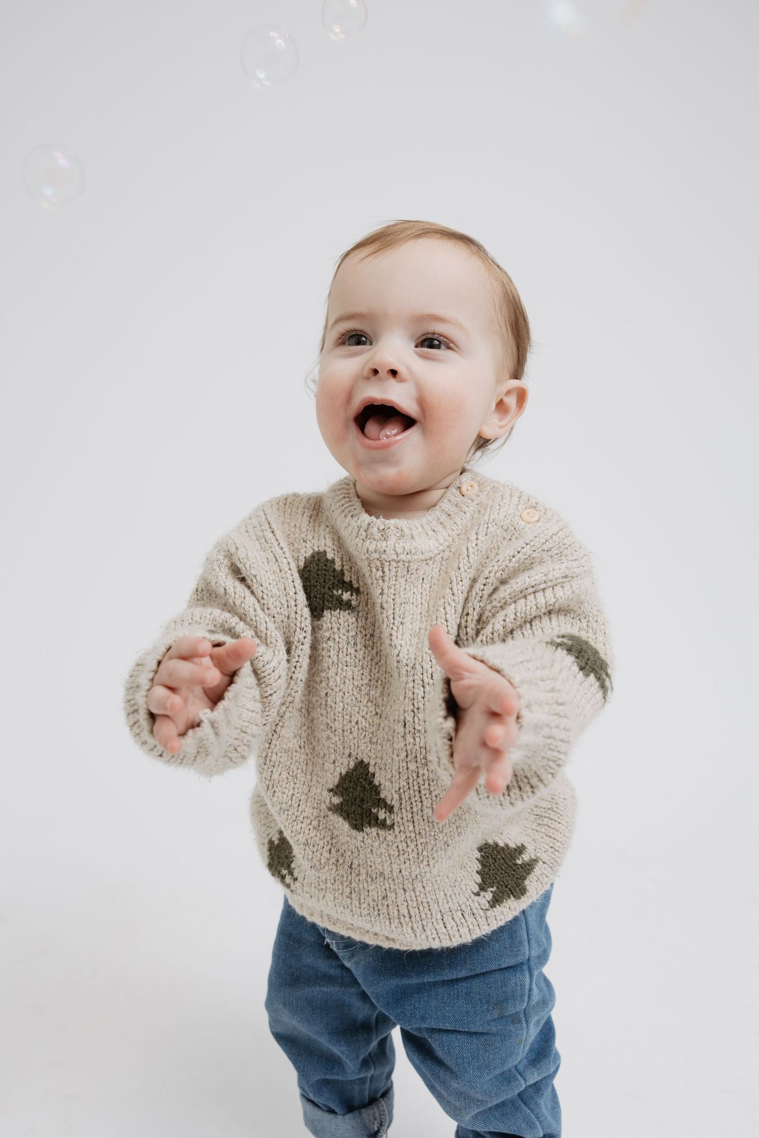 Joyful baby wearing a beige sweater with tree designs and blue jeans dances and plays against a white background.