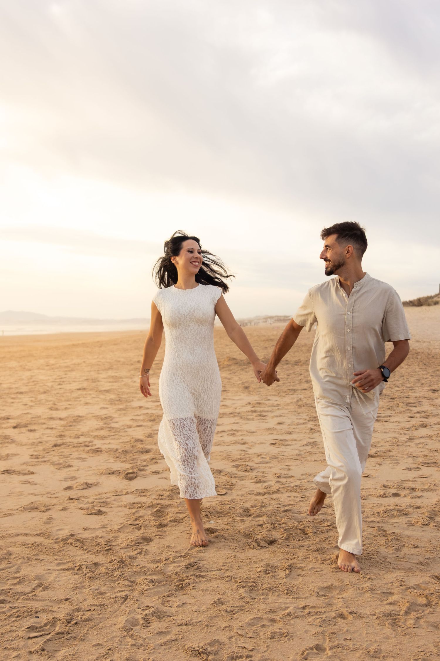 Couple in white outfits walking hand in hand on a sandy beach at sunset.