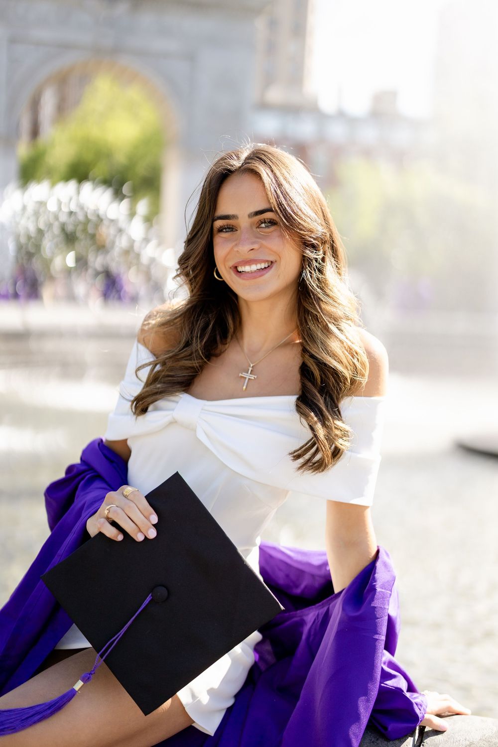 Graduate wearing purple graduation gown holds black diploma cover while smiling outdoors in sunlight.