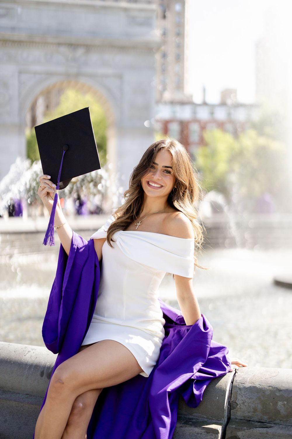 Graduate in white top and purple gown poses outdoors during graduation photoshoot.