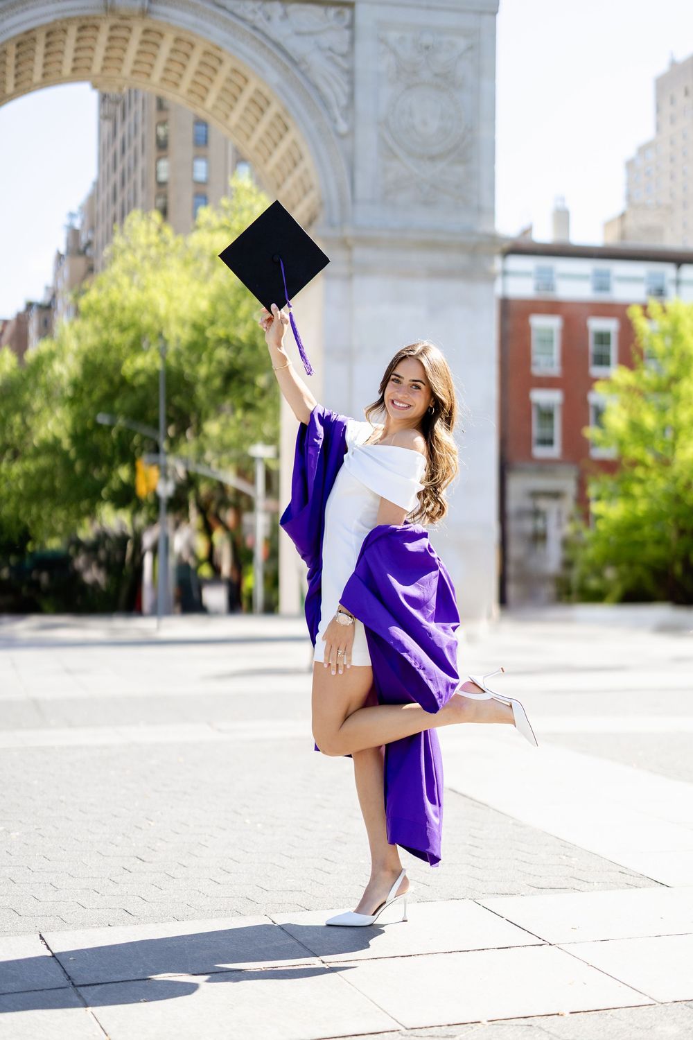 A graduate in purple gown and cap poses playfully in front of Washington Square Arch on a sunny day.