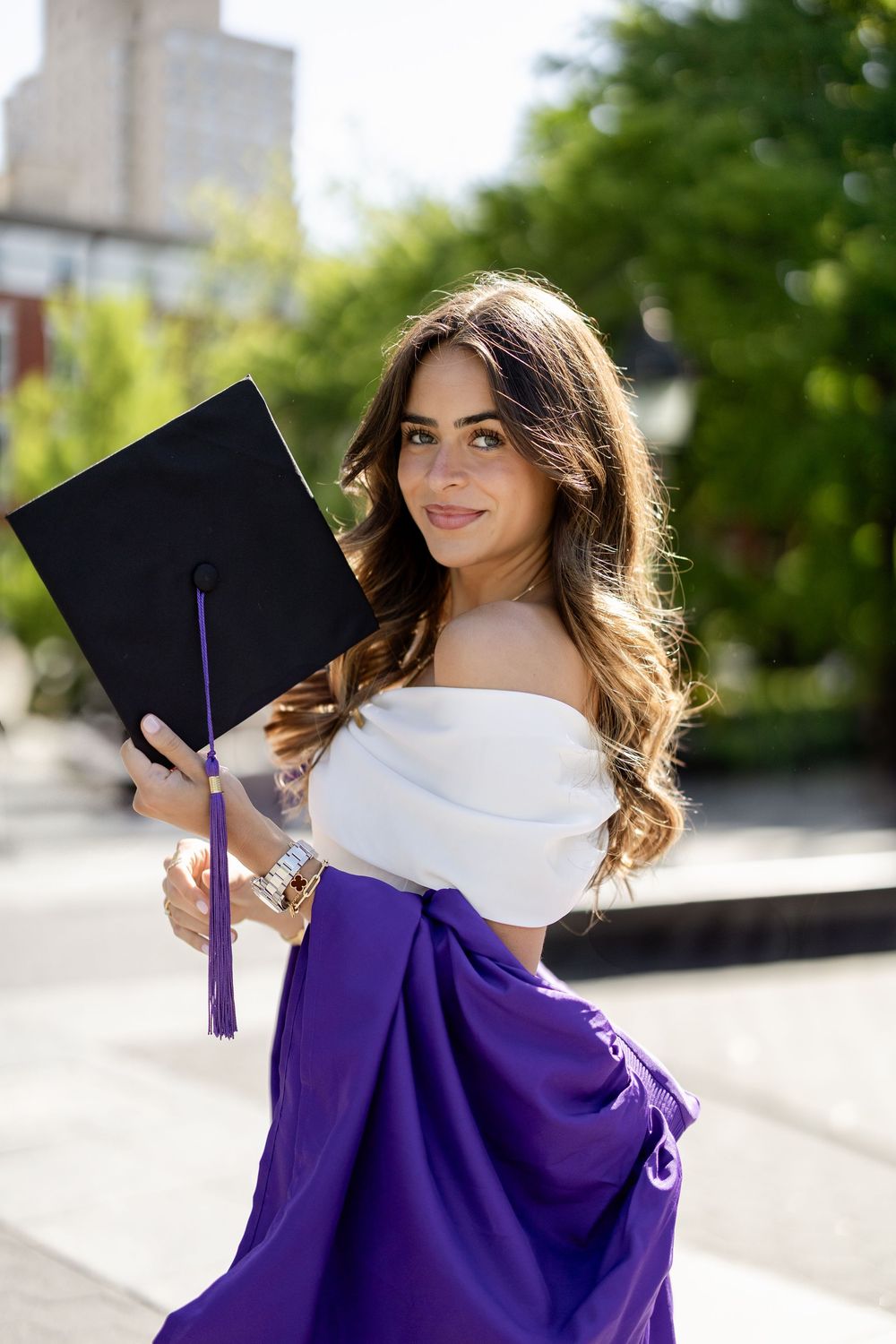 Graduate in purple cap and gown poses outdoors with wavy brown hair and white dress for graduation photos.