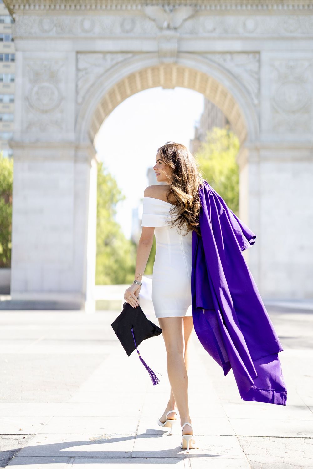 Graduate in white dress holding purple graduation gown and cap poses under ornate stone archway on sunny day.