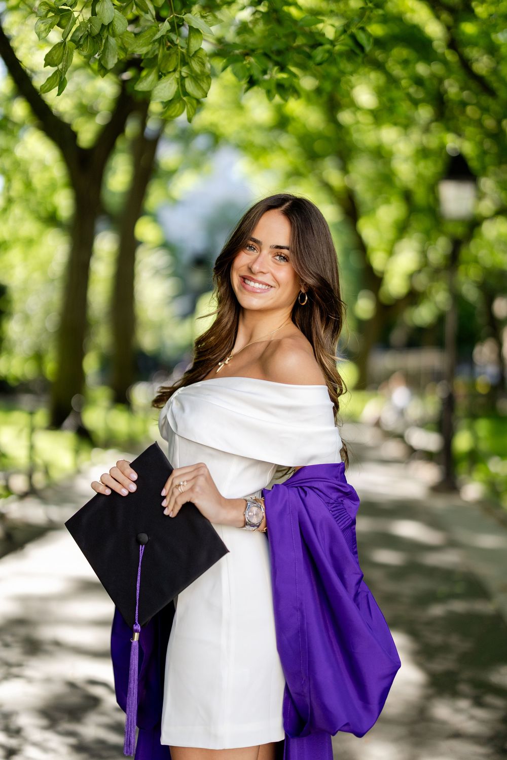 Graduate in white dress and purple graduation gown poses outdoors in a park setting with natural lighting.