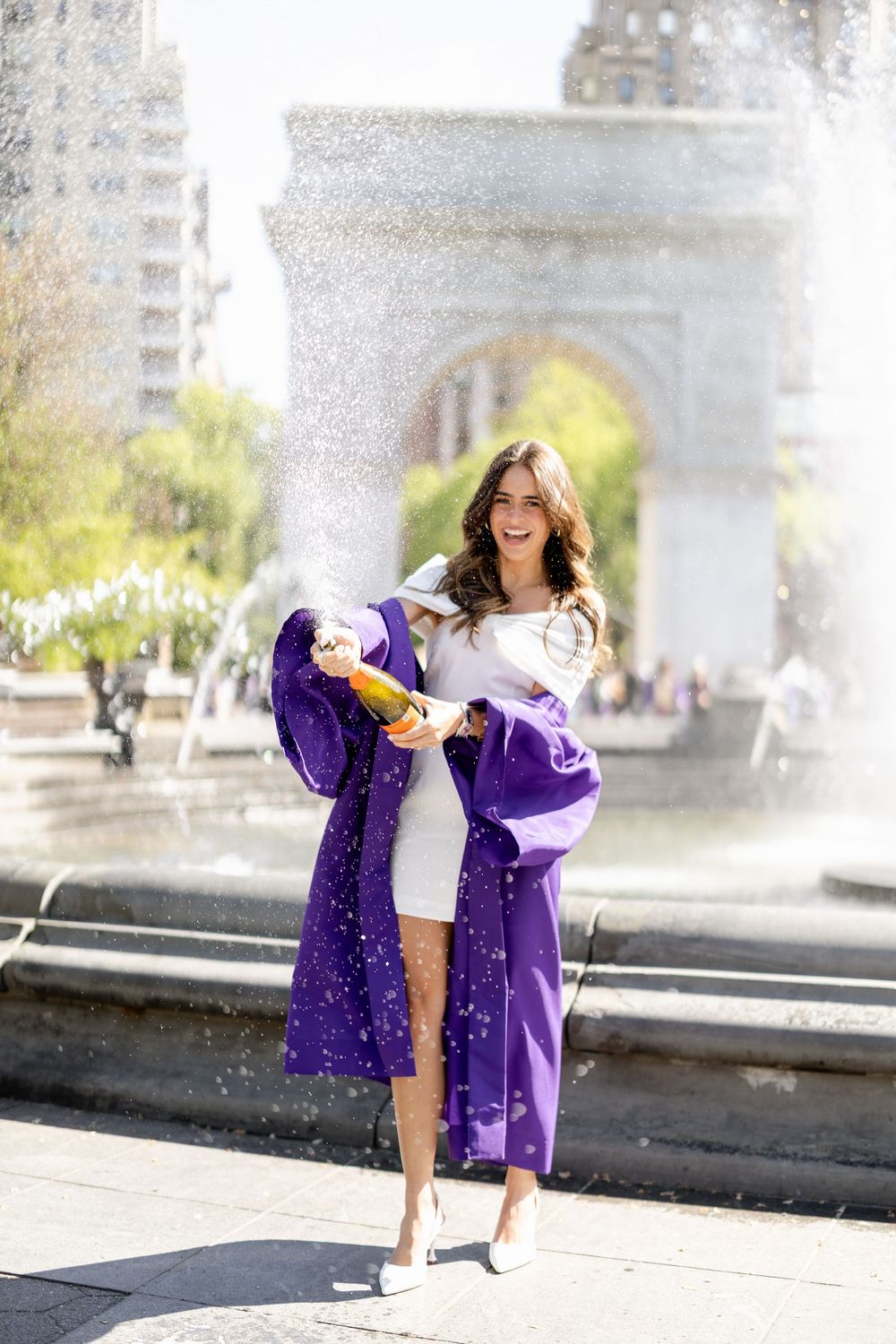 A person in a white dress and purple jacket poses playfully by a fountain in a city square on a sunny day.