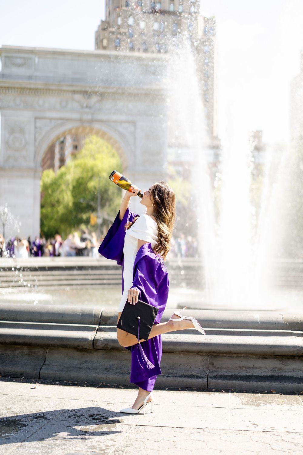 Person in purple graduation attire posing playfully near a grand fountain on sunny day.