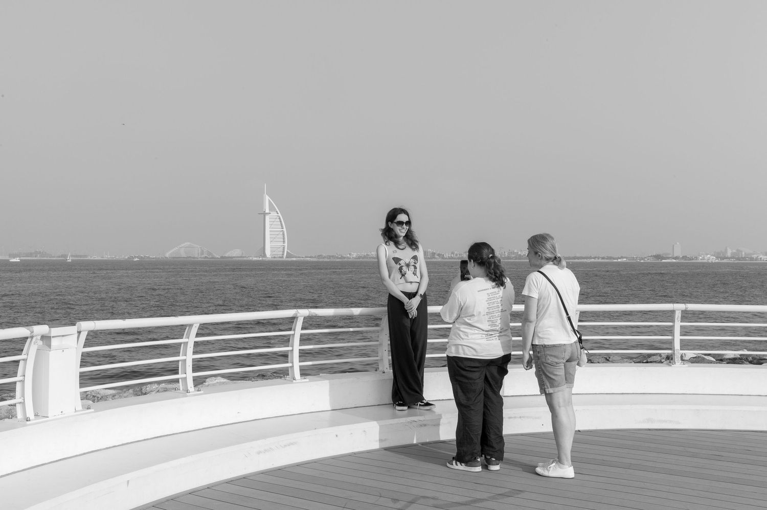 Three people stand on a cruise ship deck with Dubai's Burj Al Arab visible in the distance against a monochrome sky.