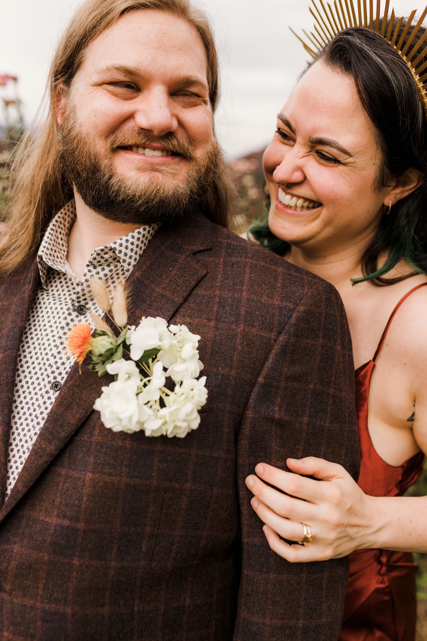 A joyful couple shares a candid moment at their wedding, with a white floral boutonniere and burgundy dress details.