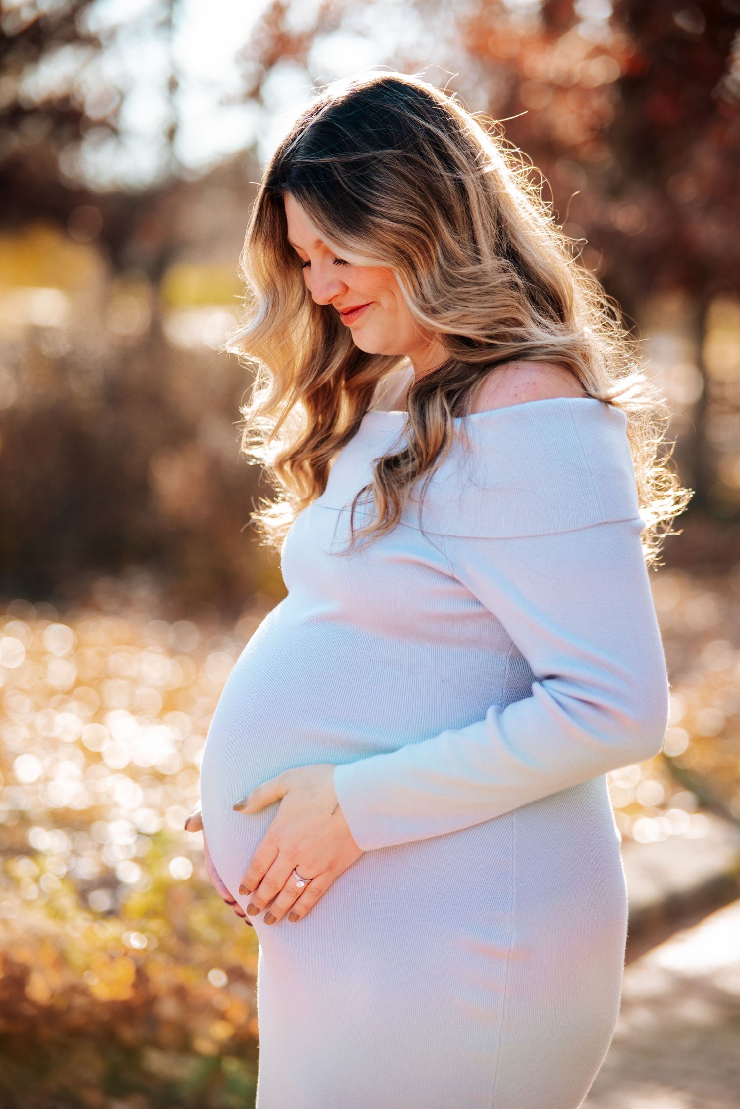 A pregnant woman in a white sweater gently cradles her baby bump during a golden hour maternity photoshoot.