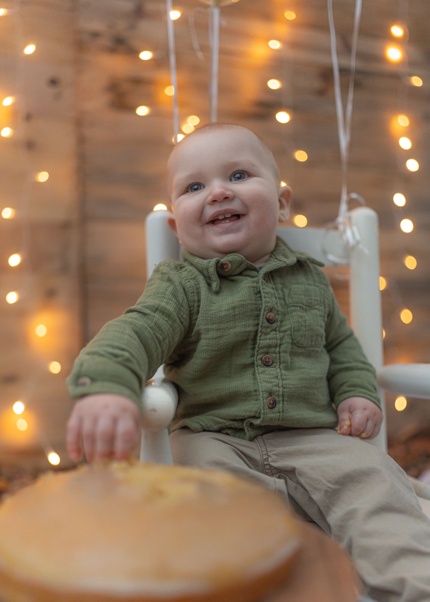 Happy baby in green button-up shirt sitting with string lights in background.