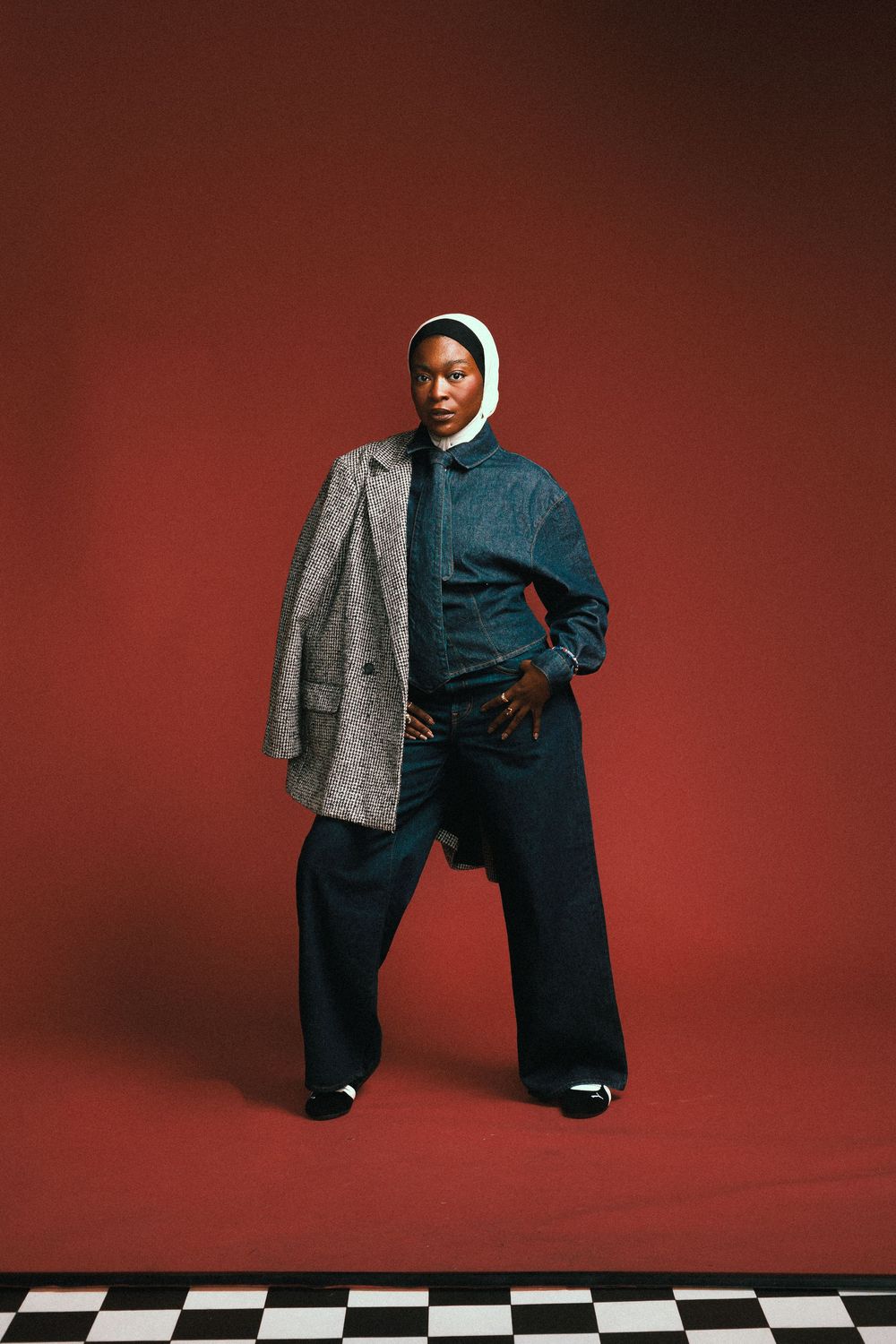 Fashion model in a white hoodie, gray blazer and wide-leg pants poses on a checkered floor against a red backdrop.