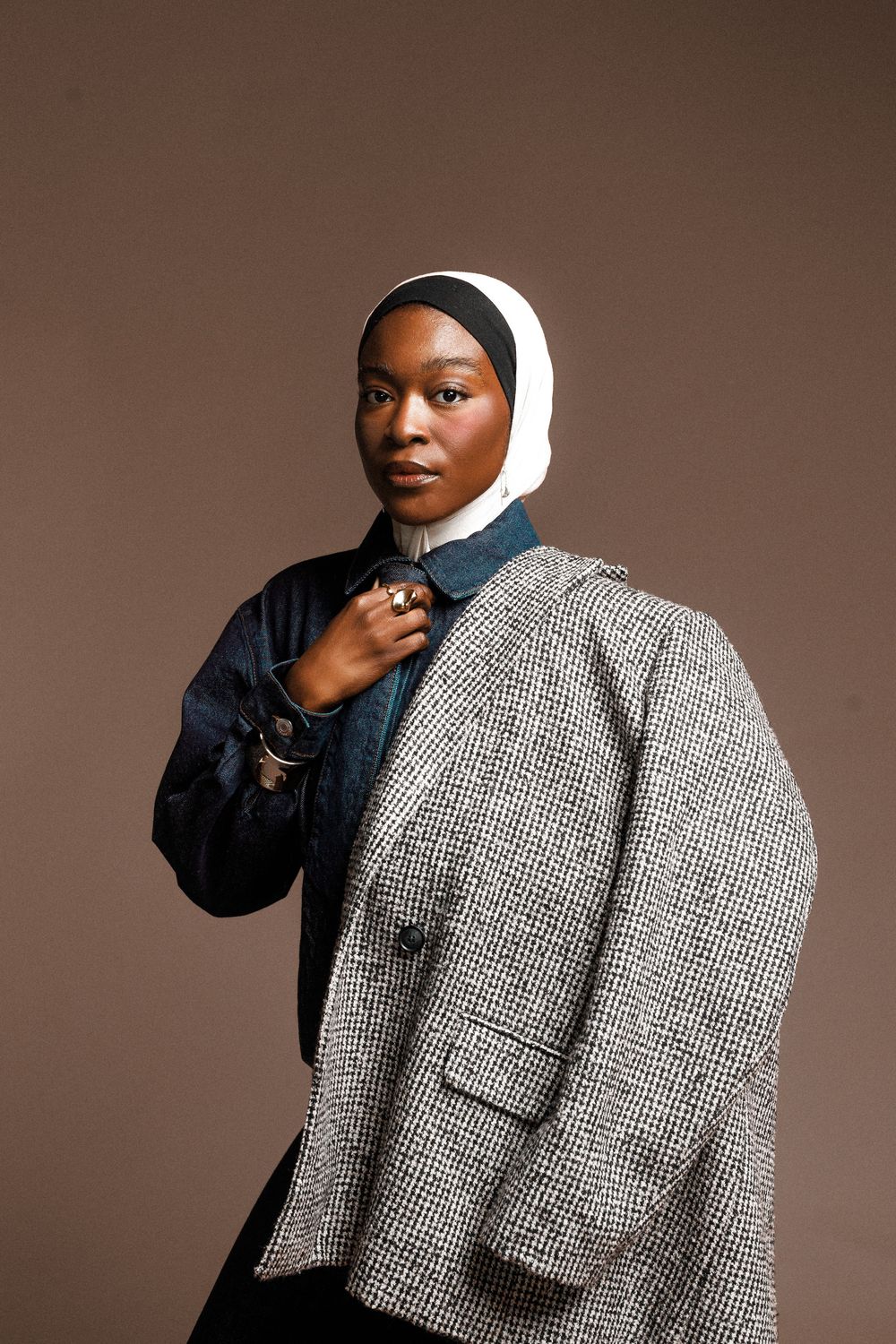 Model in white headscarf and black turtleneck poses against brown backdrop wearing textured gray tweed coat.