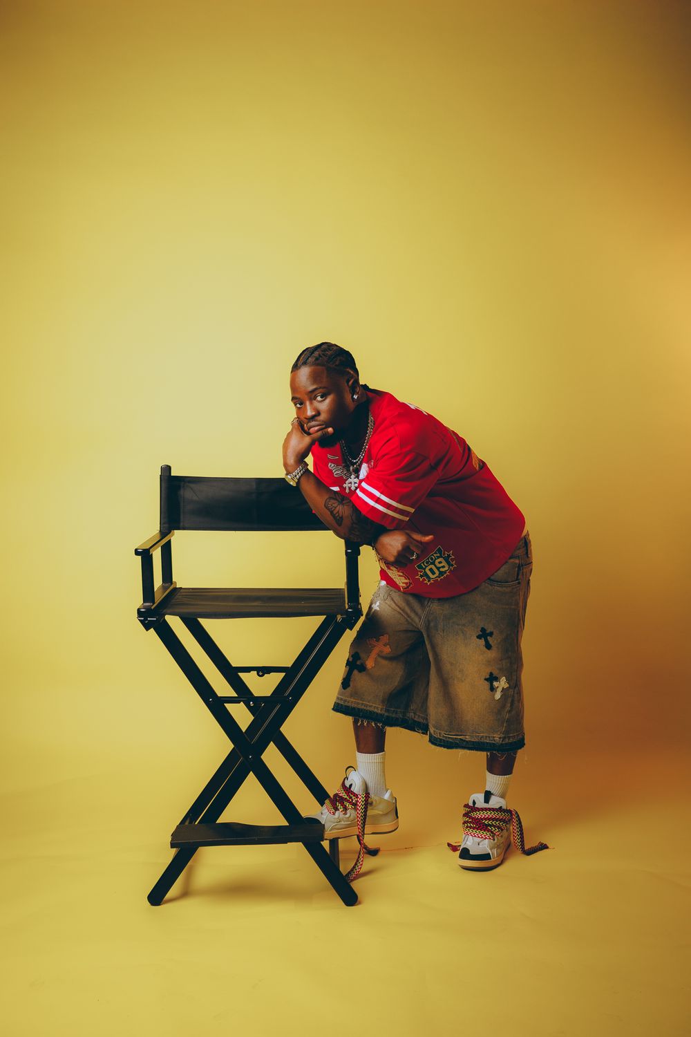 Person in a red shirt and cargo shorts leans on a black director's chair against a golden yellow backdrop in a studio setting.