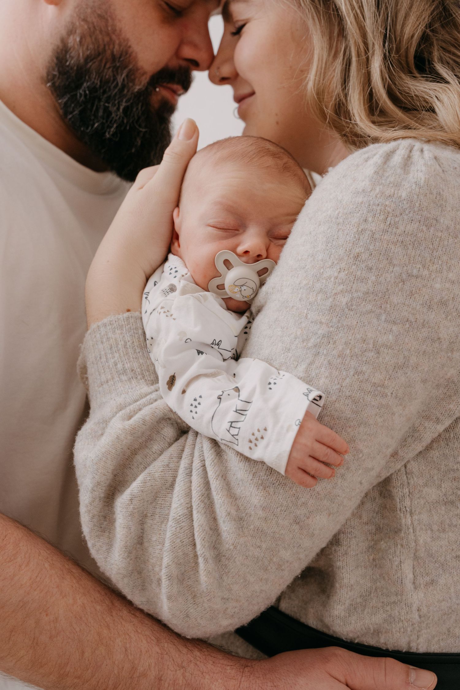 A sleeping baby with a pacifier is cradled lovingly between two adults in cozy sweaters in an intimate family portrait.