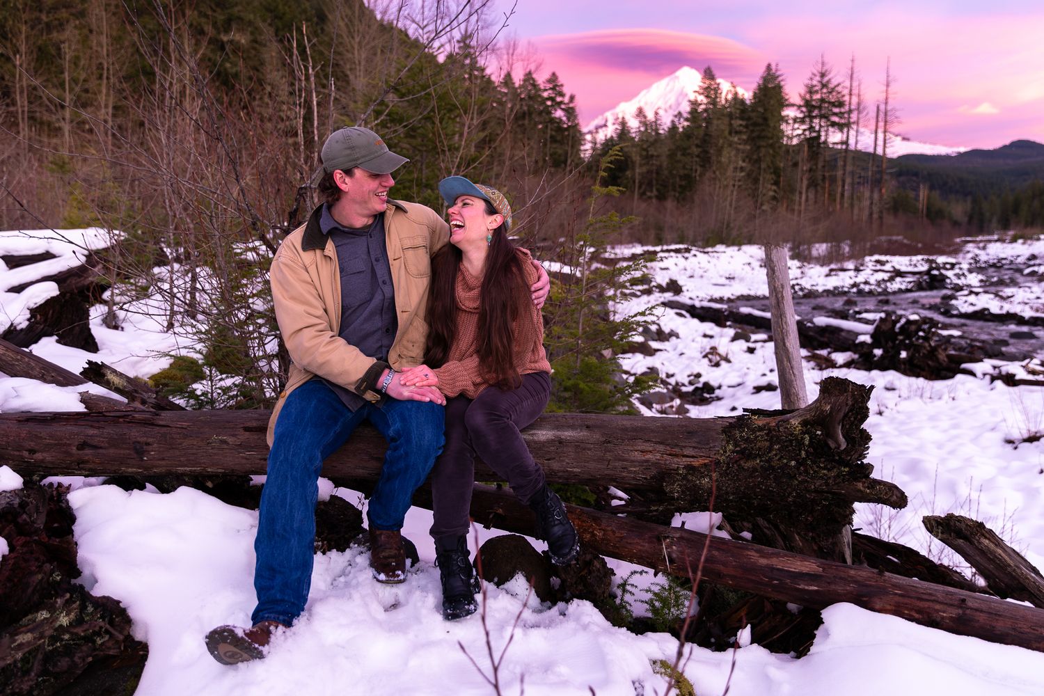 Couple sitting together on snow-covered logs in winter forest with pink sunset sky on Mount Hood.