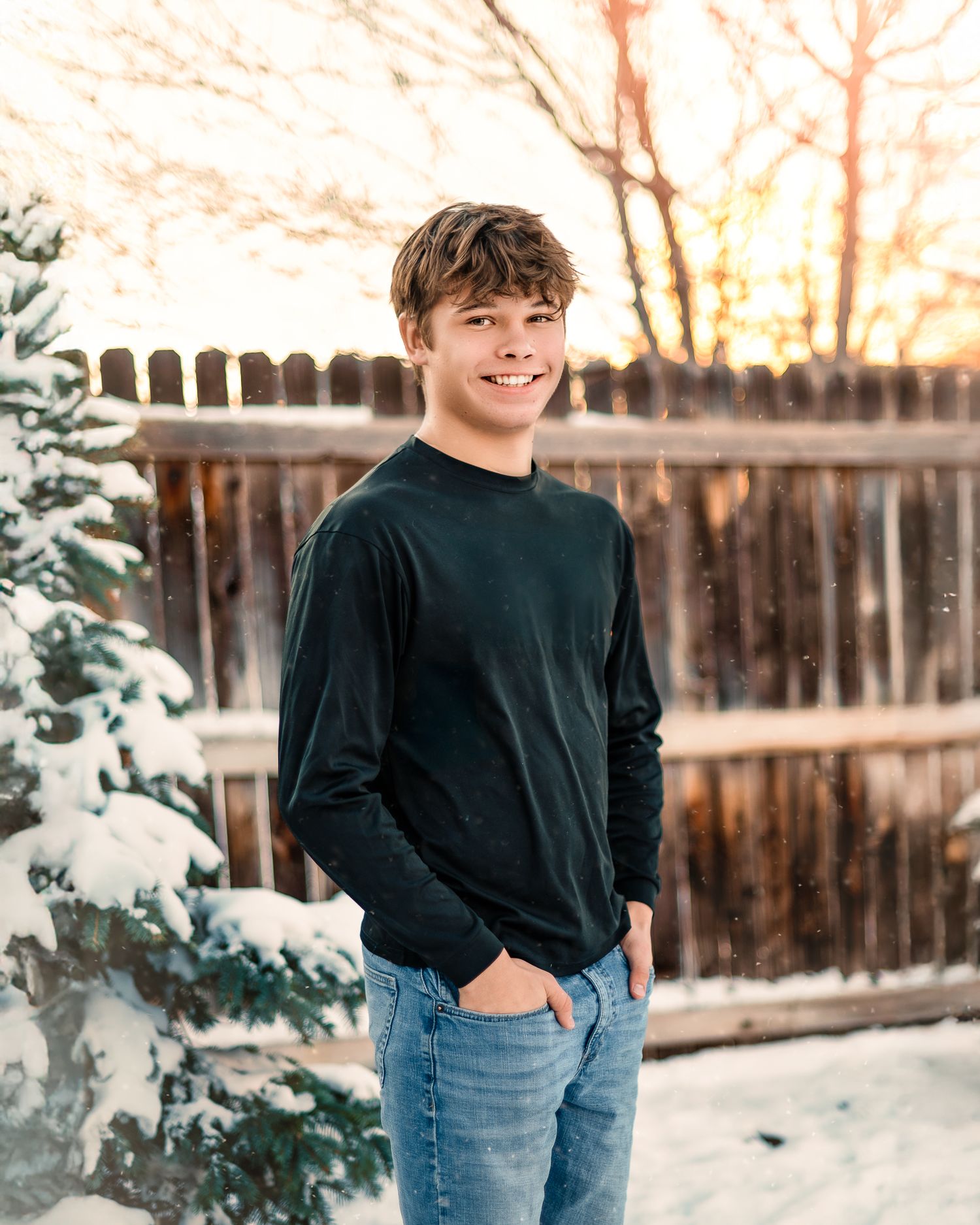 A teenage by smiling at the camera in front of a snowy evergreen tree in a Denver, CO backyard