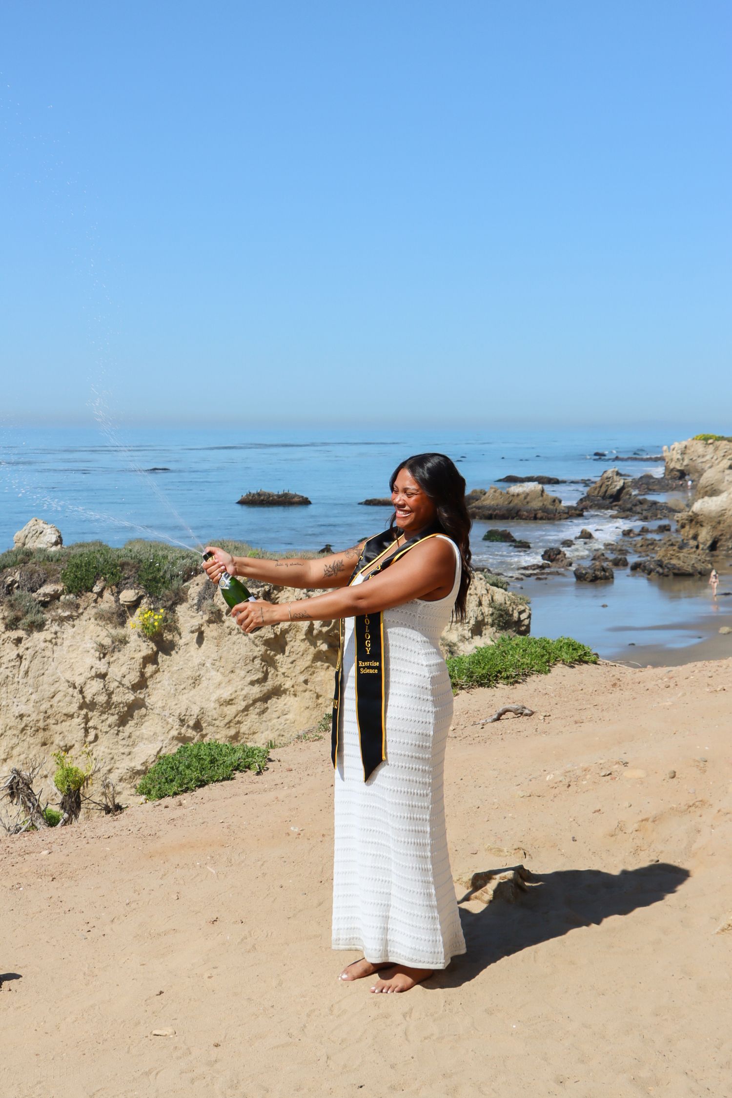 Graduate in white dress poses dramatically on rocky beach with ocean view.