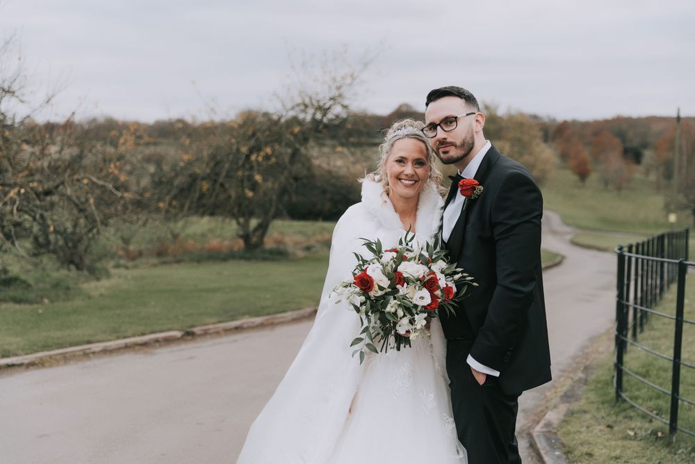 A couple in formal wedding attire walks together along a scenic outdoor path during an autumn day.