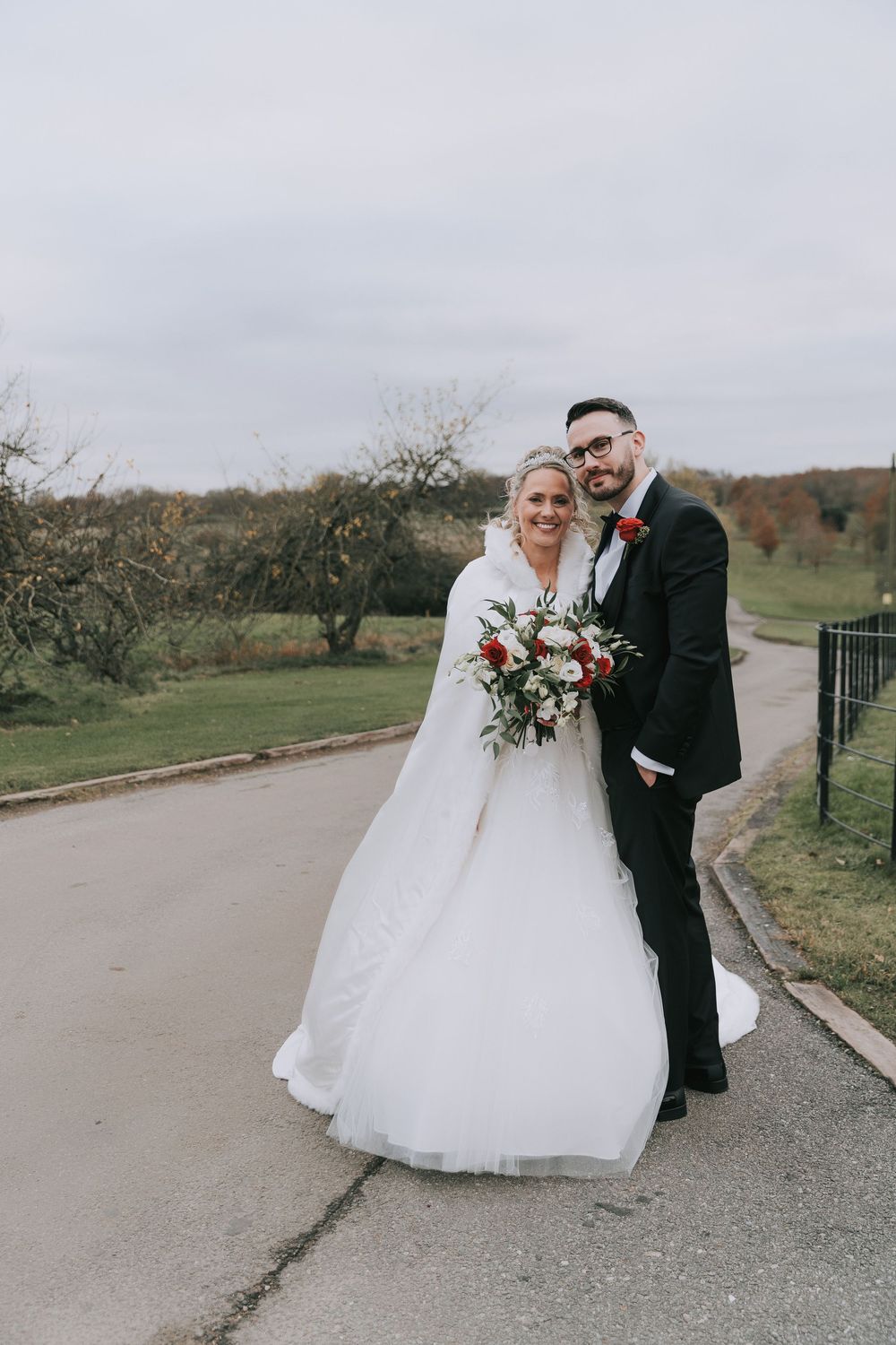 A couple poses together on a winding path during their autumn wedding celebration, surrounded by trees and greenery.