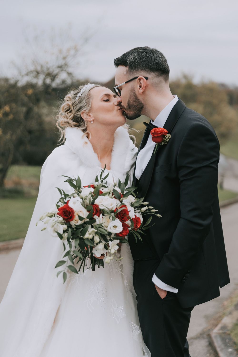 A romantic wedding couple sharing a kiss outdoors while holding a bouquet with red and white flowers.
