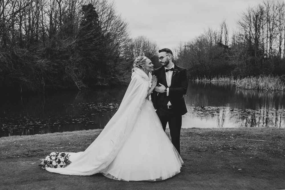 Romantic wedding couple poses in a serene winter landscape with bare trees and a tranquil pond in the background.