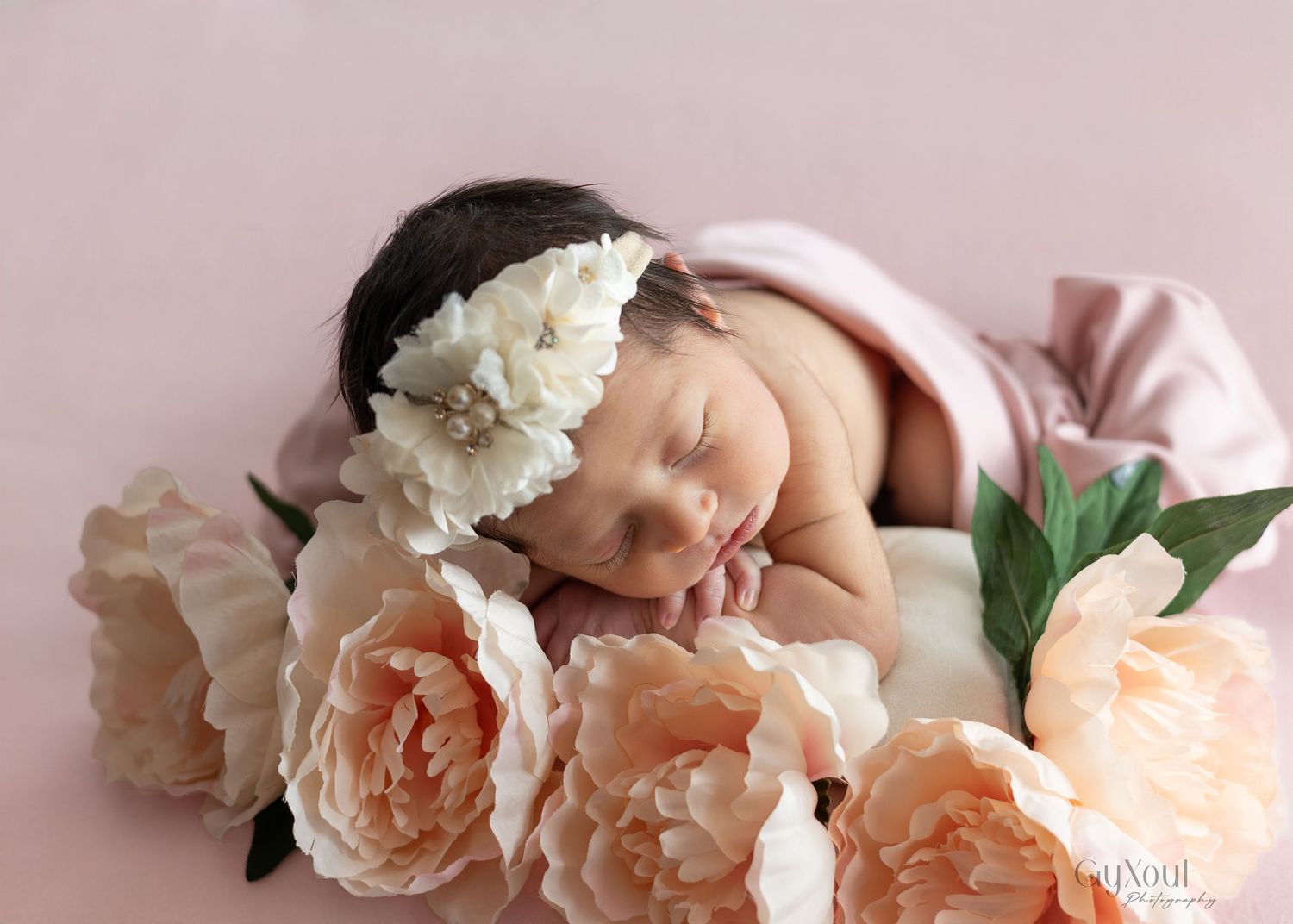 Peaceful sleeping newborn nestled among soft peach roses and white flowers on pink background.