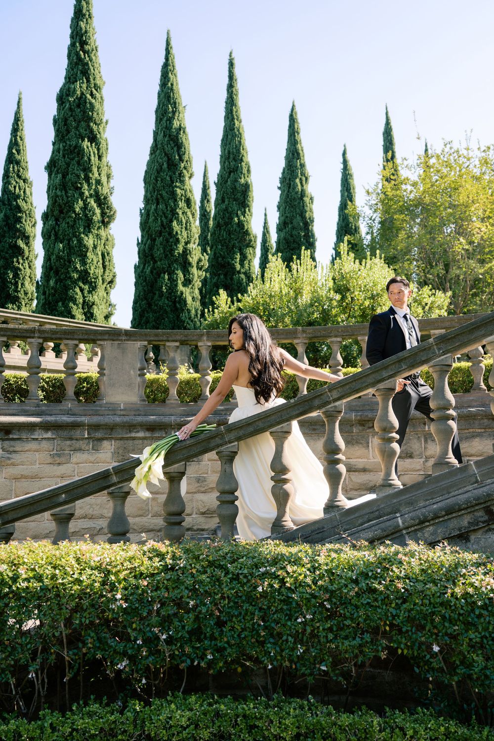 A couple poses on an elegant stone terrace with Italian cypress trees and ornate balustrades in the background.