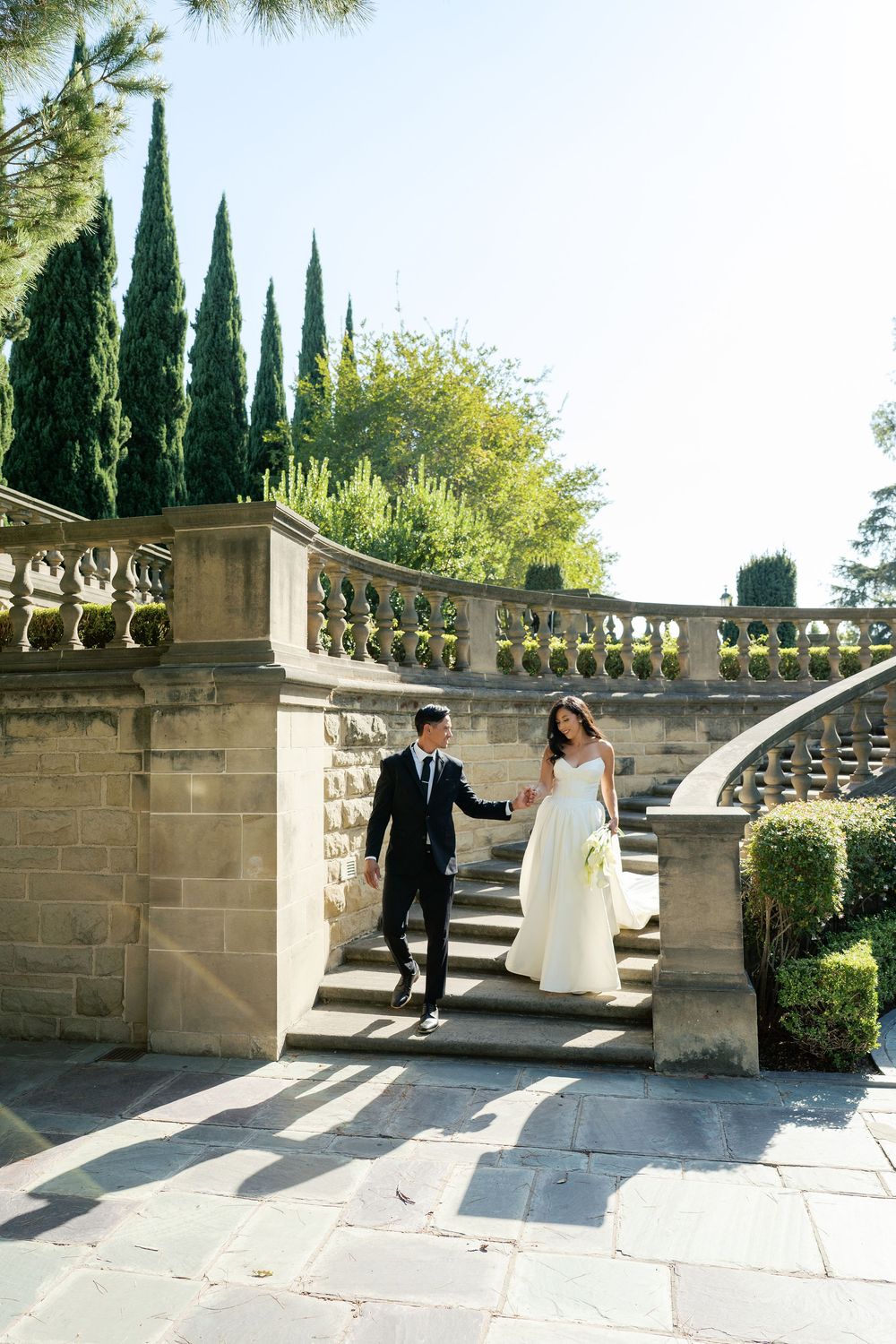 Wedding couple walking along a grand stone staircase surrounded by cypress trees and classical architecture.