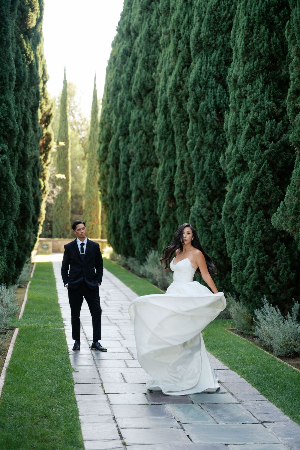 A couple poses on a stone path lined with tall cypress trees, with the bride's white dress flowing in the breeze.