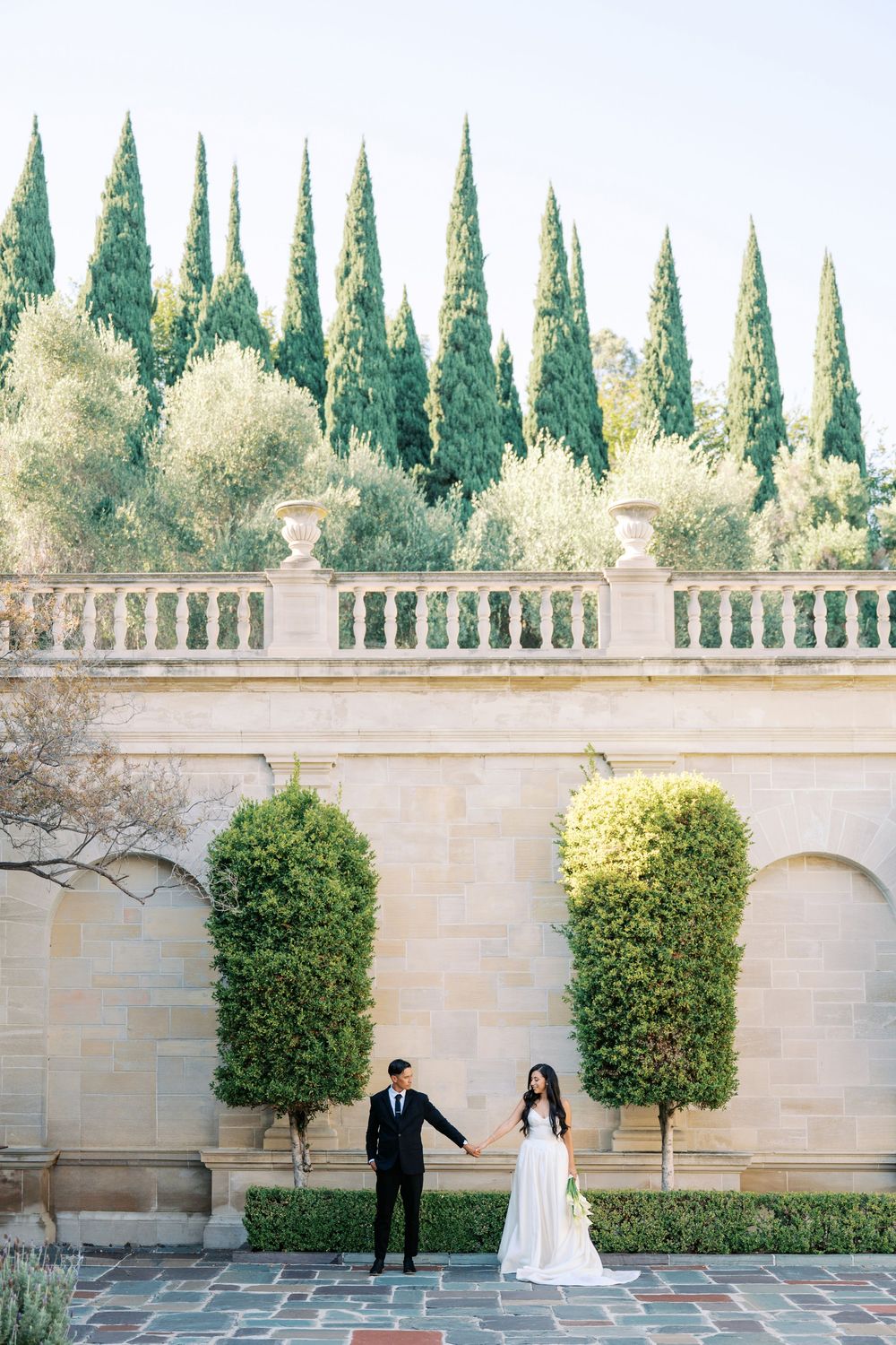 Romantic wedding photo shoot in front of a stone wall with manicured topiary and cypress trees.