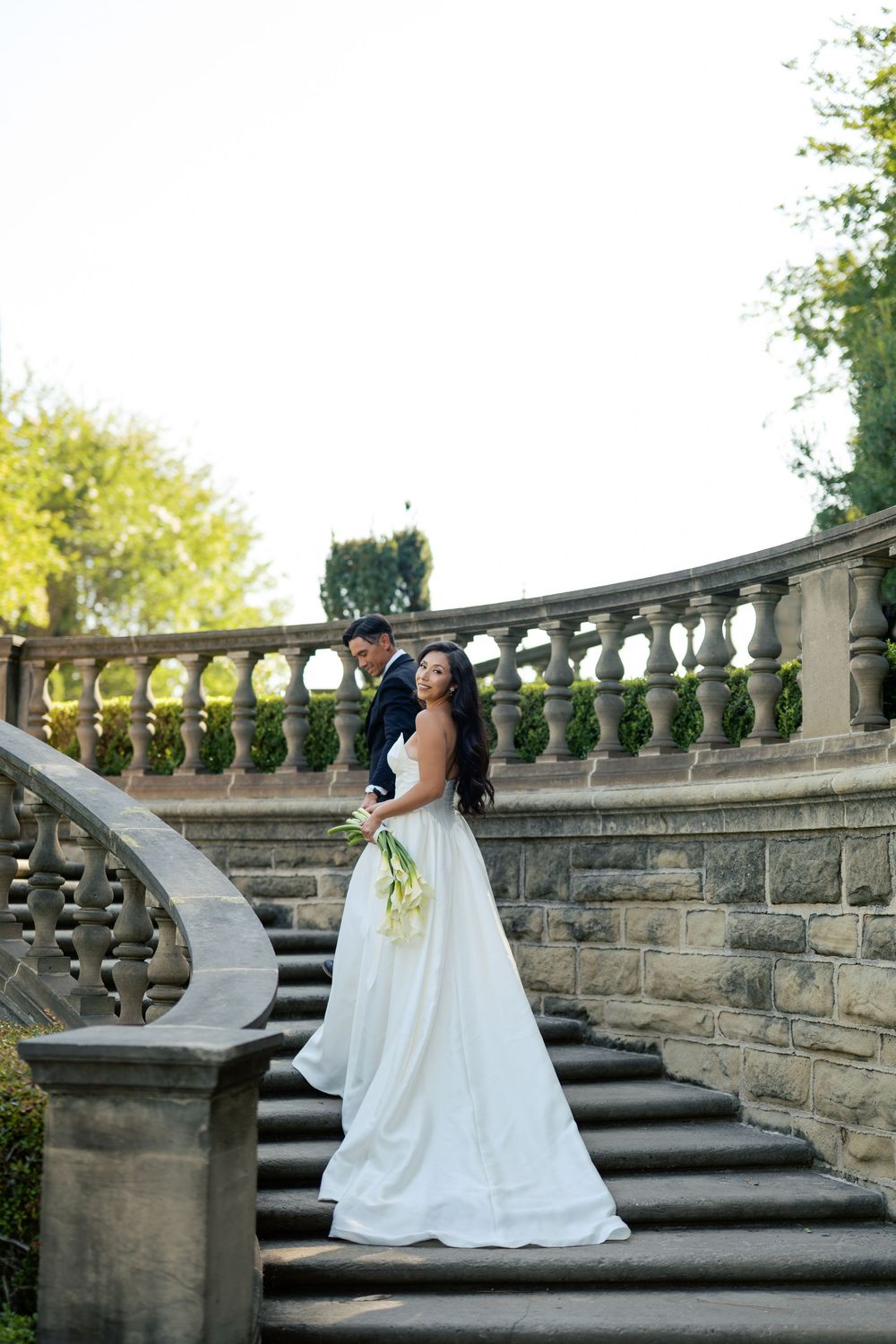 A bride in a flowing white gown poses on elegant stone stairs with ornate balustrades in a garden setting.