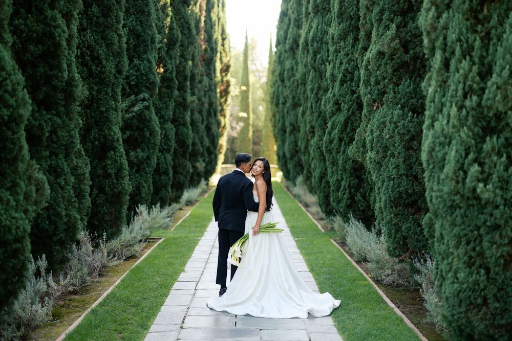 A couple in wedding attire share a romantic moment on a path lined with tall cypress trees in a formal garden.