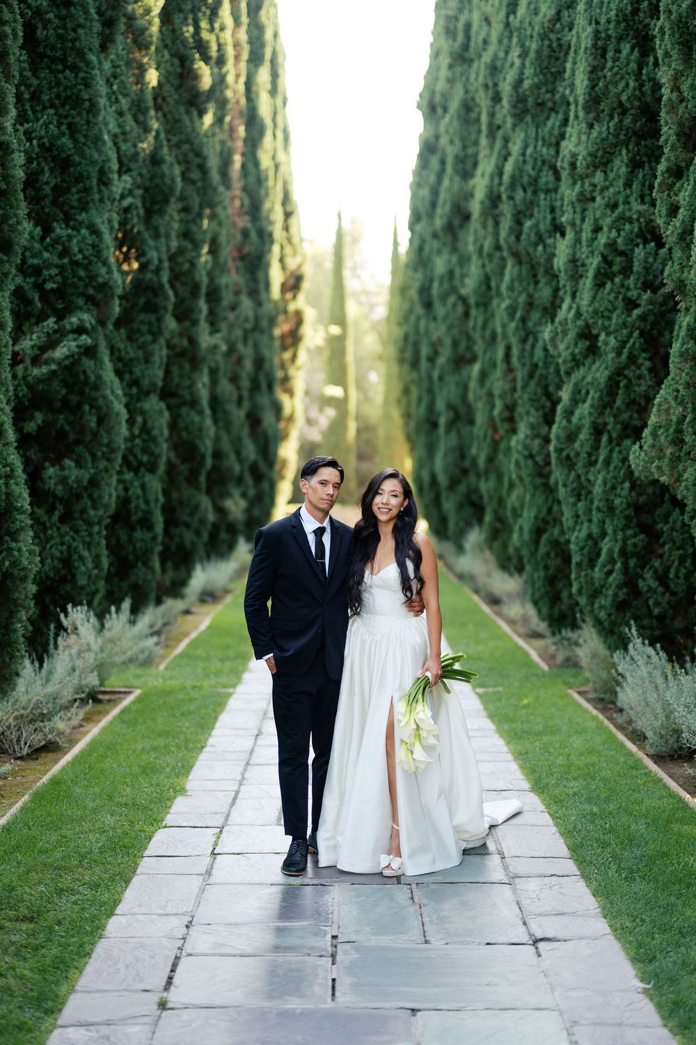 A couple stands together on a garden path flanked by towering cypress trees creating a dramatic perspective.