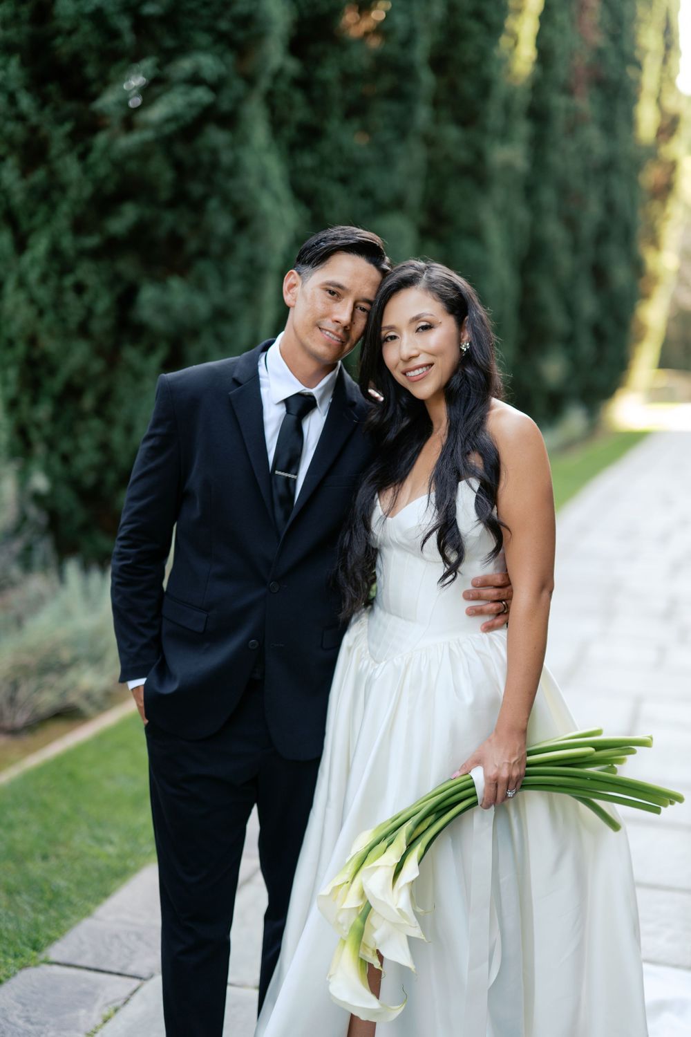 Elegant wedding couple poses together with calla lily bouquet against a backdrop of cypress trees.