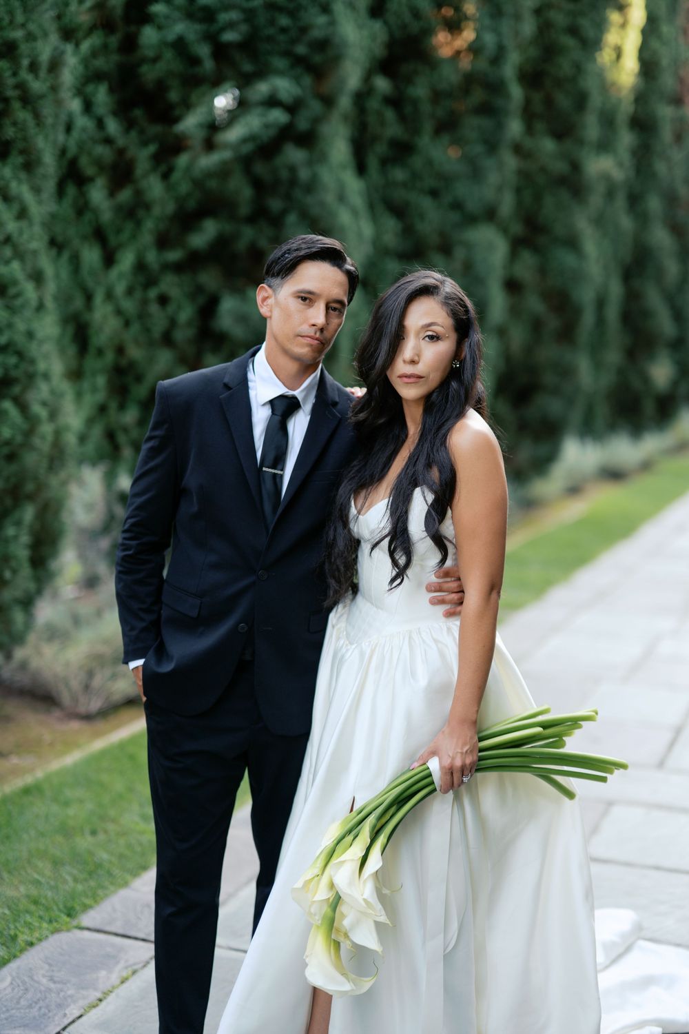 A couple in wedding attire poses together against a green garden backdrop with white calla lily bouquet.
