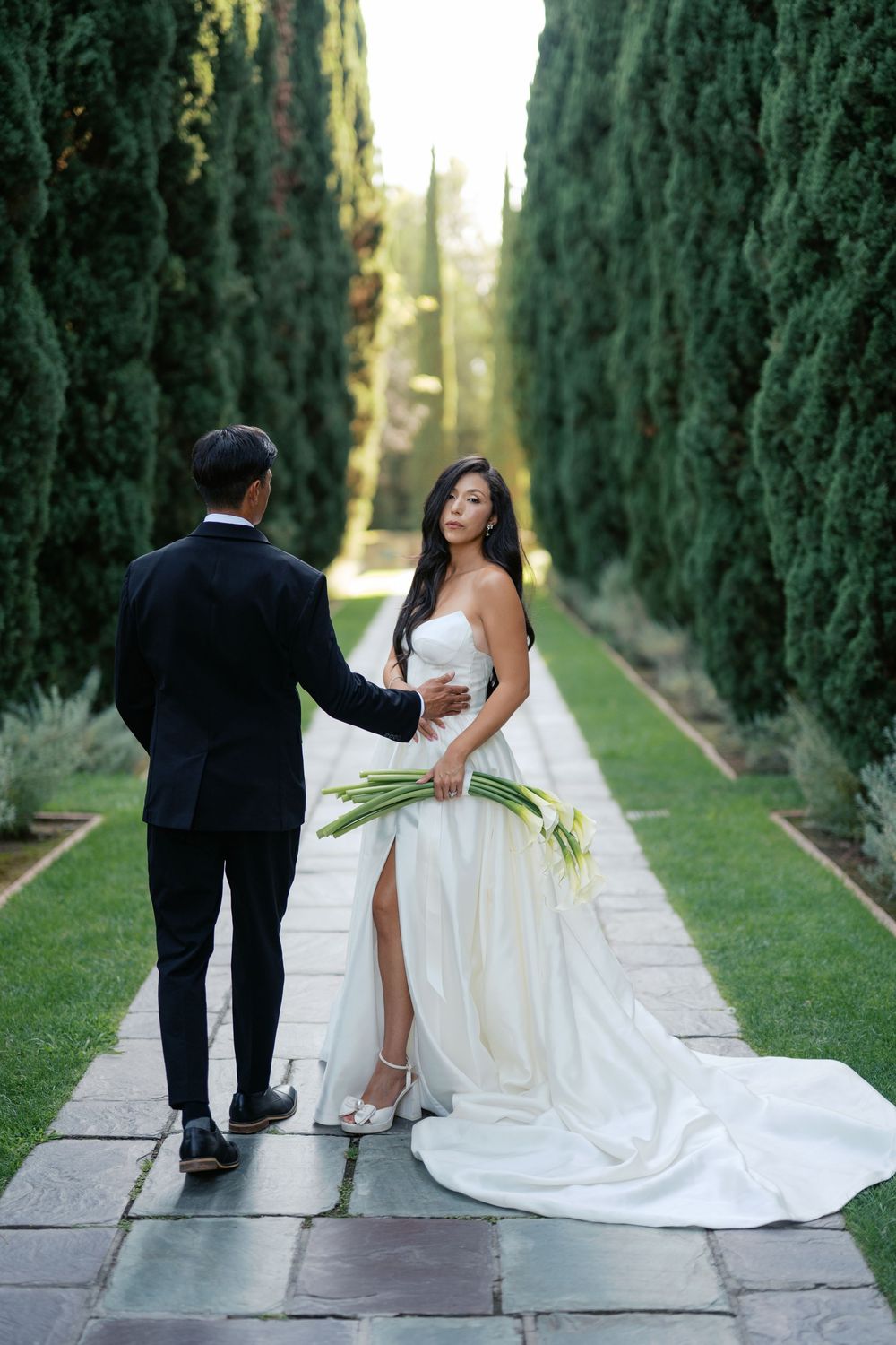 Newlyweds pose playfully on stone pathway surrounded by tall cypress trees.