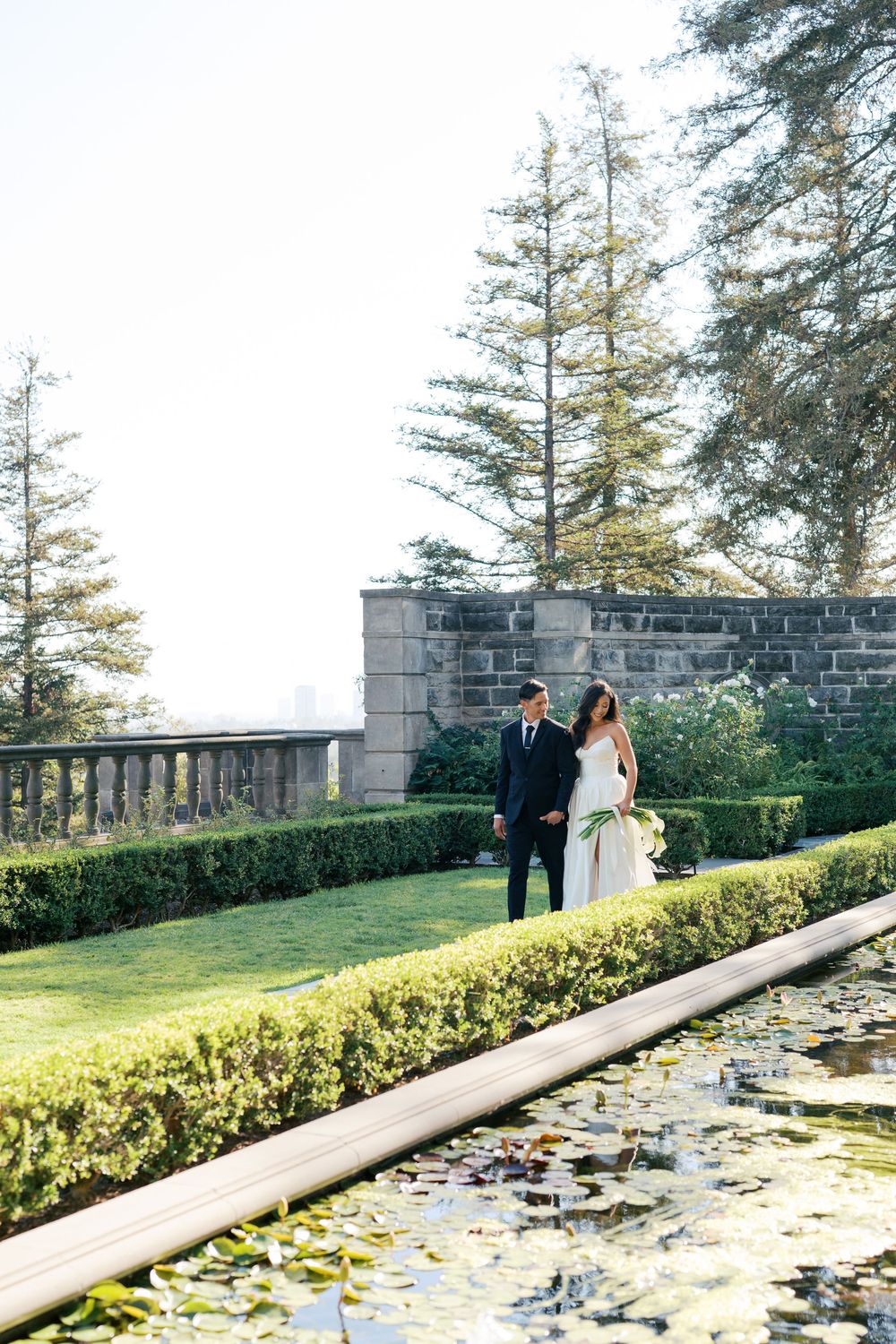Couple in formal attire walks along stone wall with hedges and evergreen trees in background.