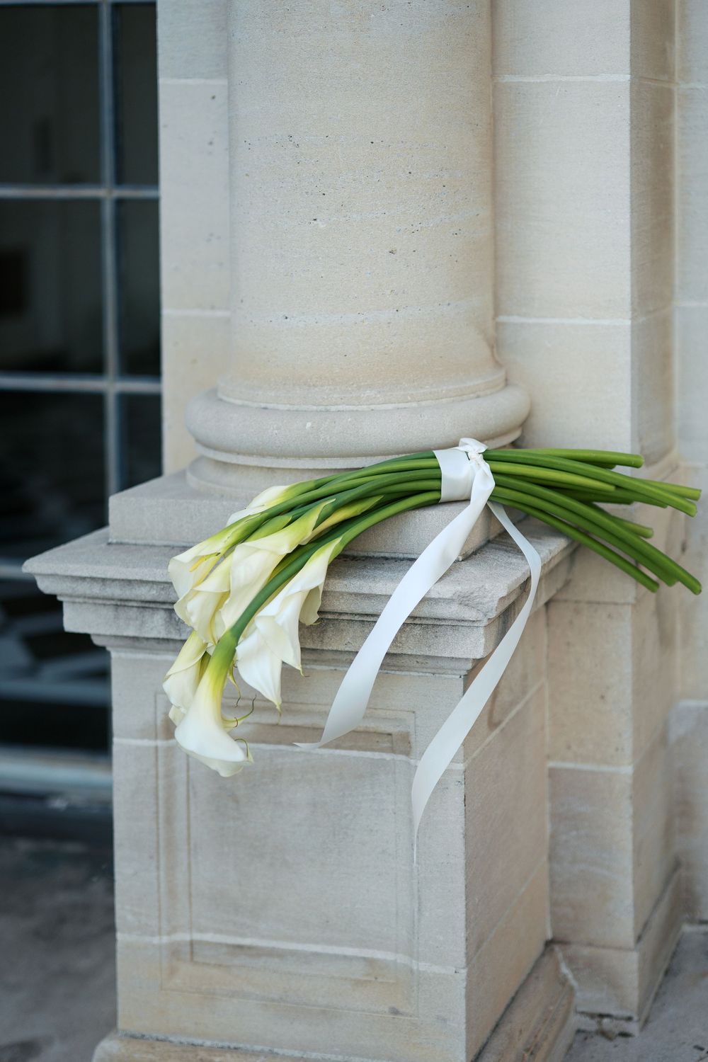 White calla lily bouquet with ribbons rests on a stone column ledge.
